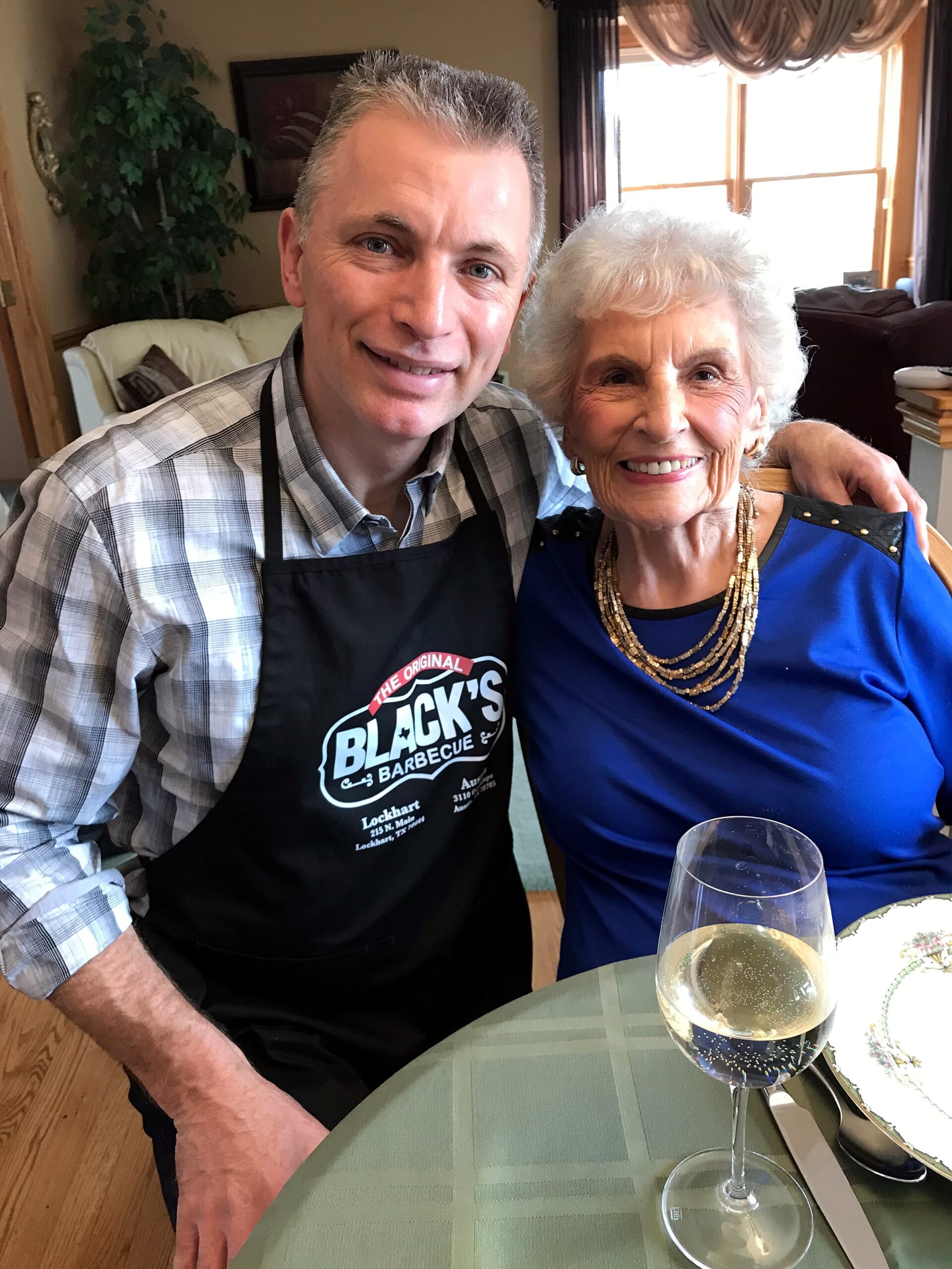 Smiling man in a plaid shirt wearing a Black's Barbecue apron sits beside an elderly woman in a blue dress at a dining table, with a glass of white wine in the foreground.