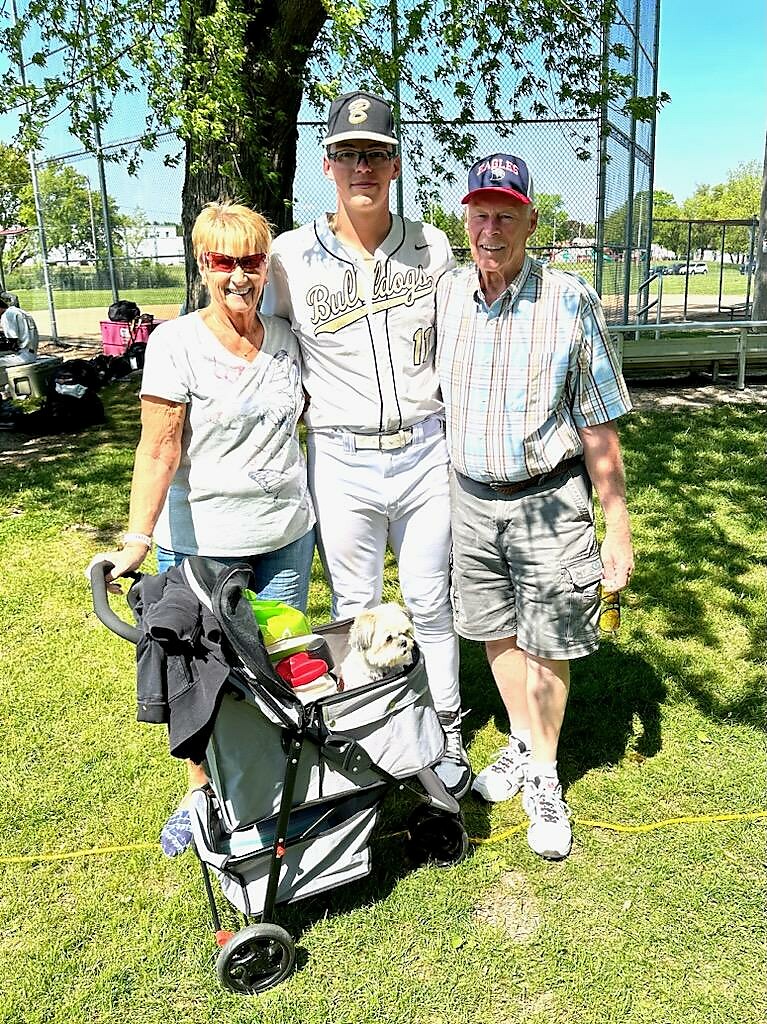 Young baseball player in Bulldogs uniform (number 11) stands with a smiling woman and man on a sunny field; a small dog sits in a wagon full of gear.