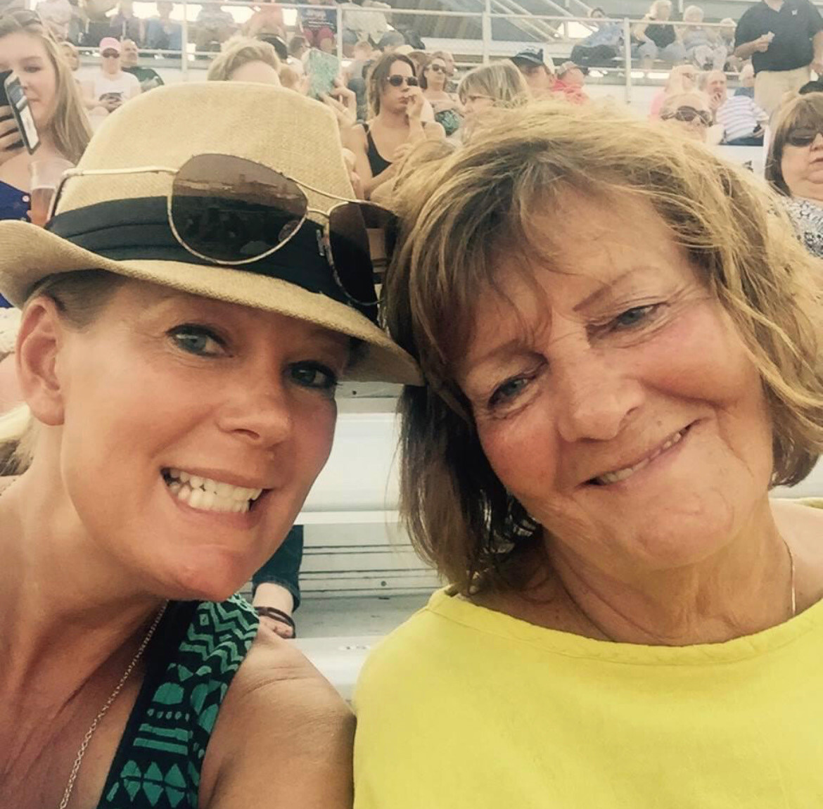 Two women close together, smiling at the camera at a crowded outdoor event; one wears a tan hat with sunglasses on it.