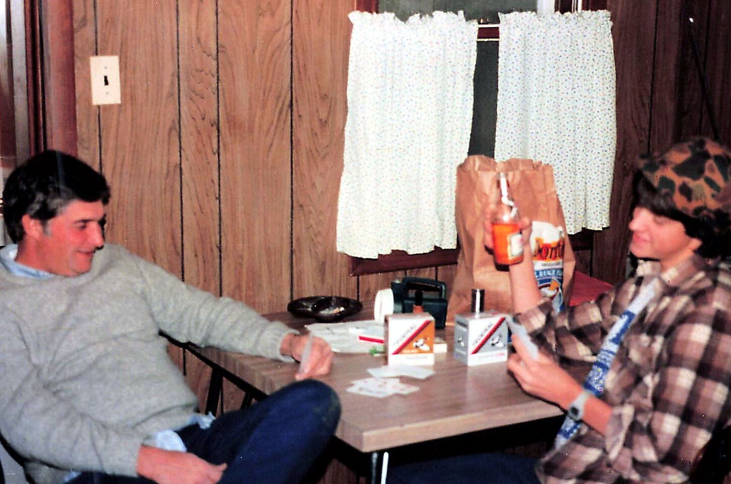 Two friends sitting at a wood-paneled table playing cards; one raises a beer bottle for a toast.