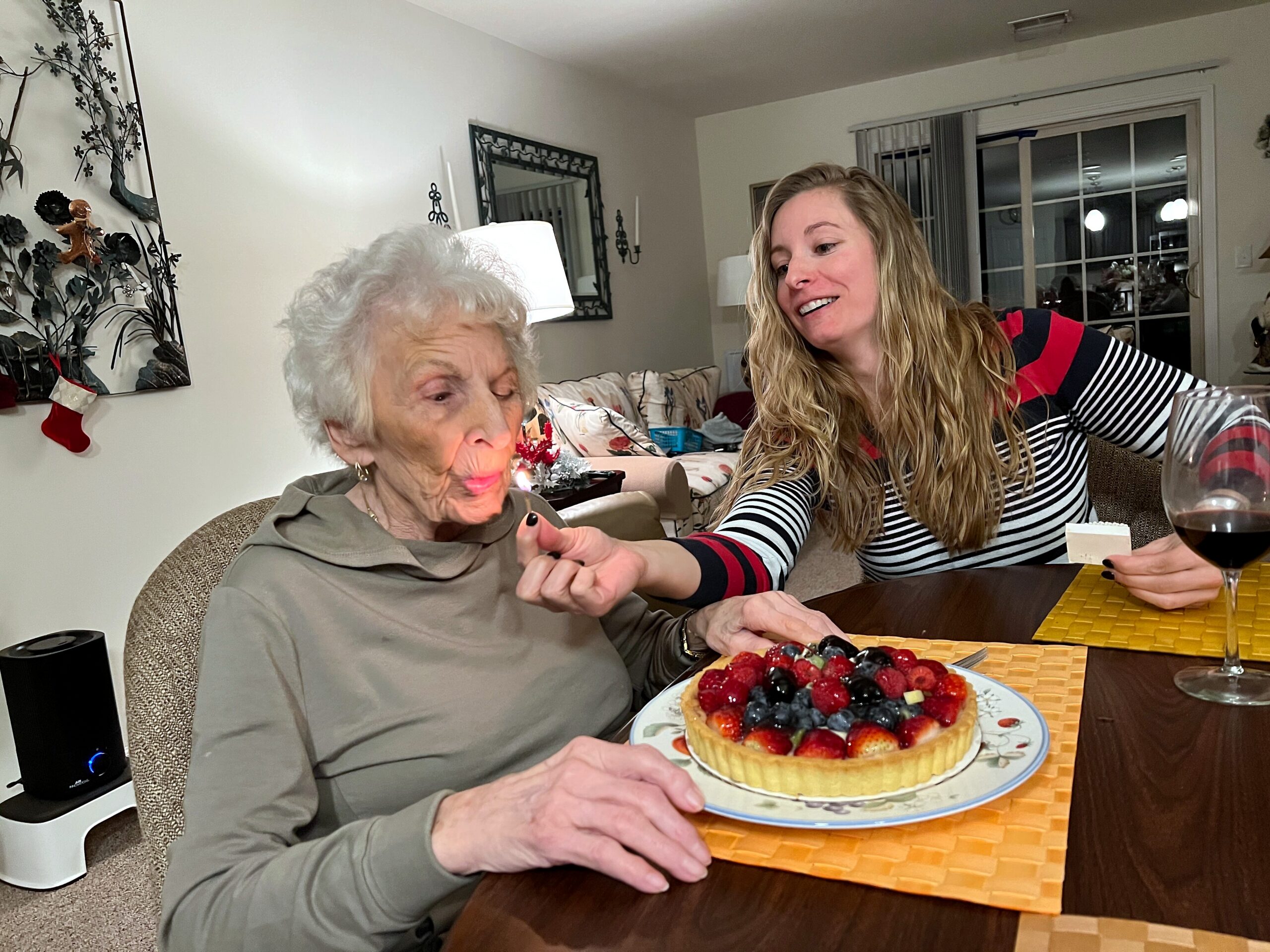 Older woman being fed berries by a smiling younger woman at a dining table, with a berry tart on a plate in the foreground.