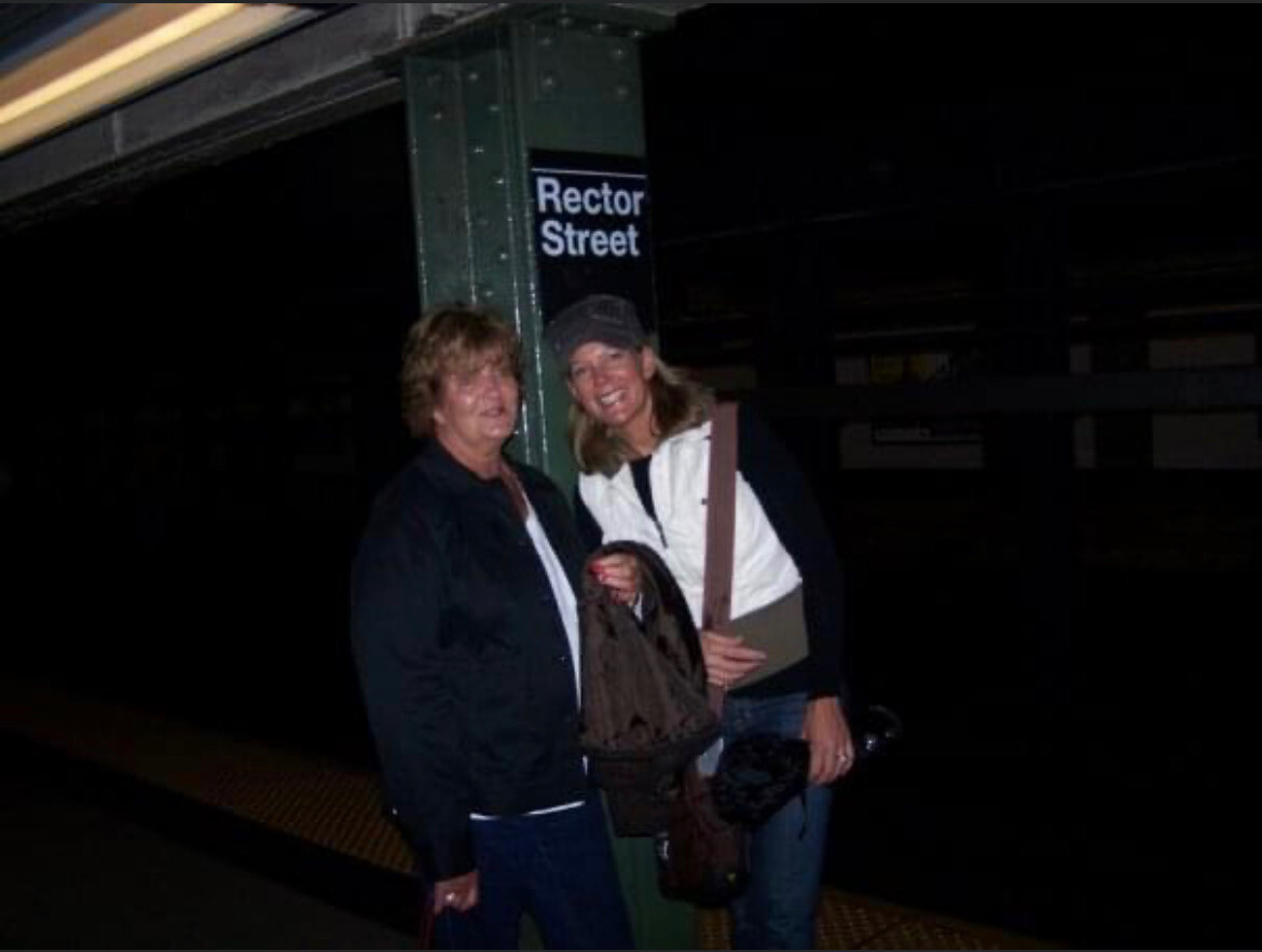 Two smiling women standing on a subway platform near a pillar with a Rector Street sign at night, carrying bags and jackets.