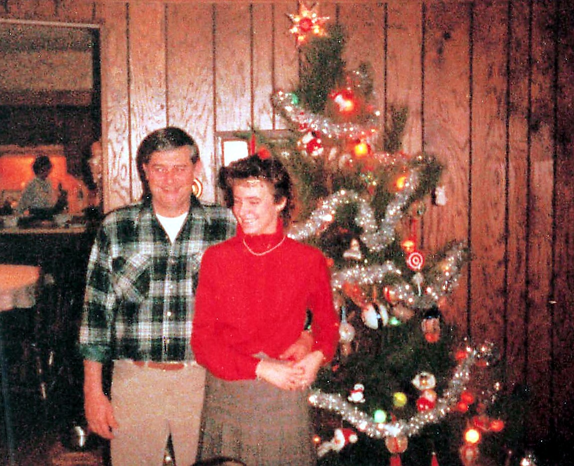 A couple stands beside a decorated Christmas tree indoors, smiling at the camera.