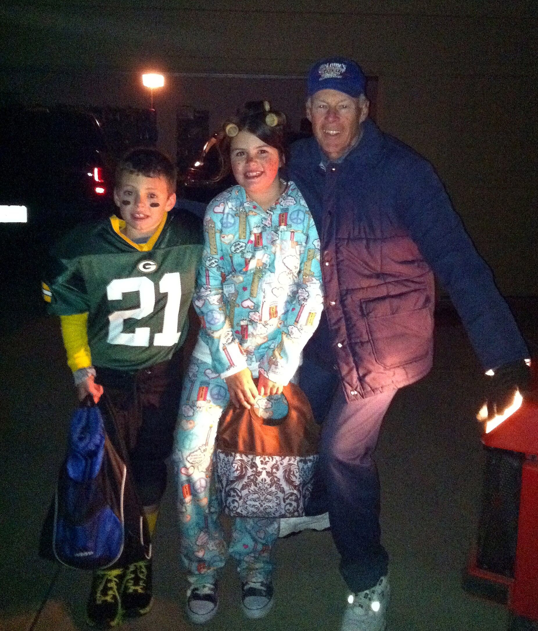 Three people posing in a dark outdoor setting: a man in a blue jacket and cap with two children in front, one in a green jersey with face paint and the other in colorful pajamas; a small fire glows on the right.