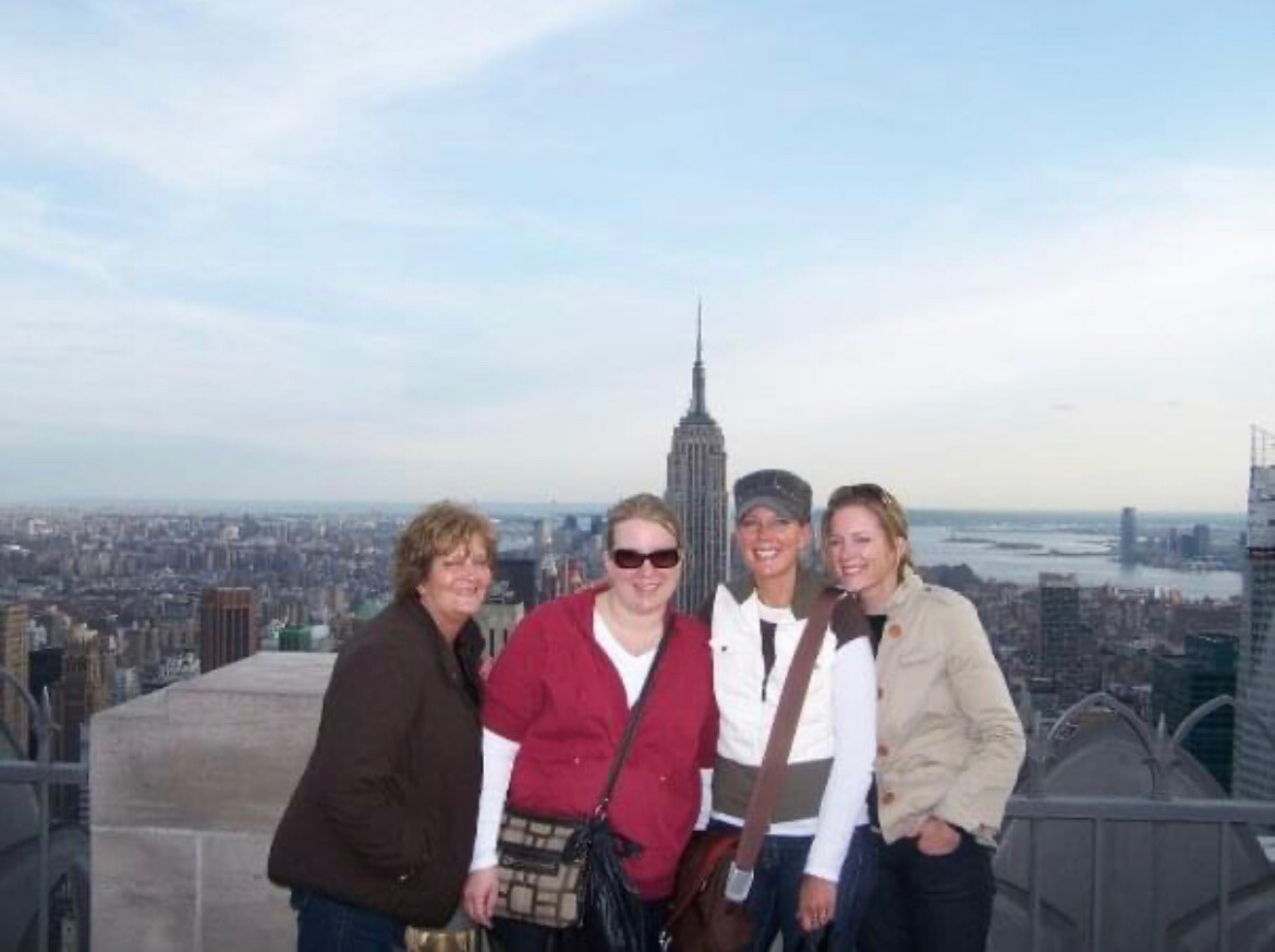 Four women posing and smiling on a rooftop with the Manhattan skyline and the Empire State Building in the background.
