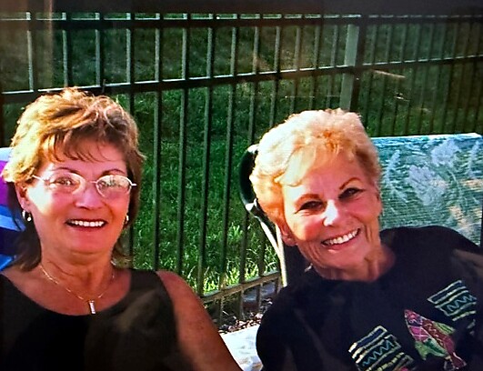 Two smiling older women sitting outdoors on a bench by a fence, enjoying a sunny day.