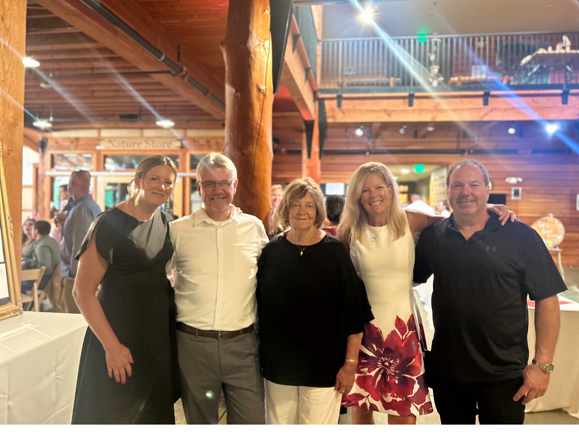 Group of five adults posing together and smiling in a warm, wood-paneled event space.