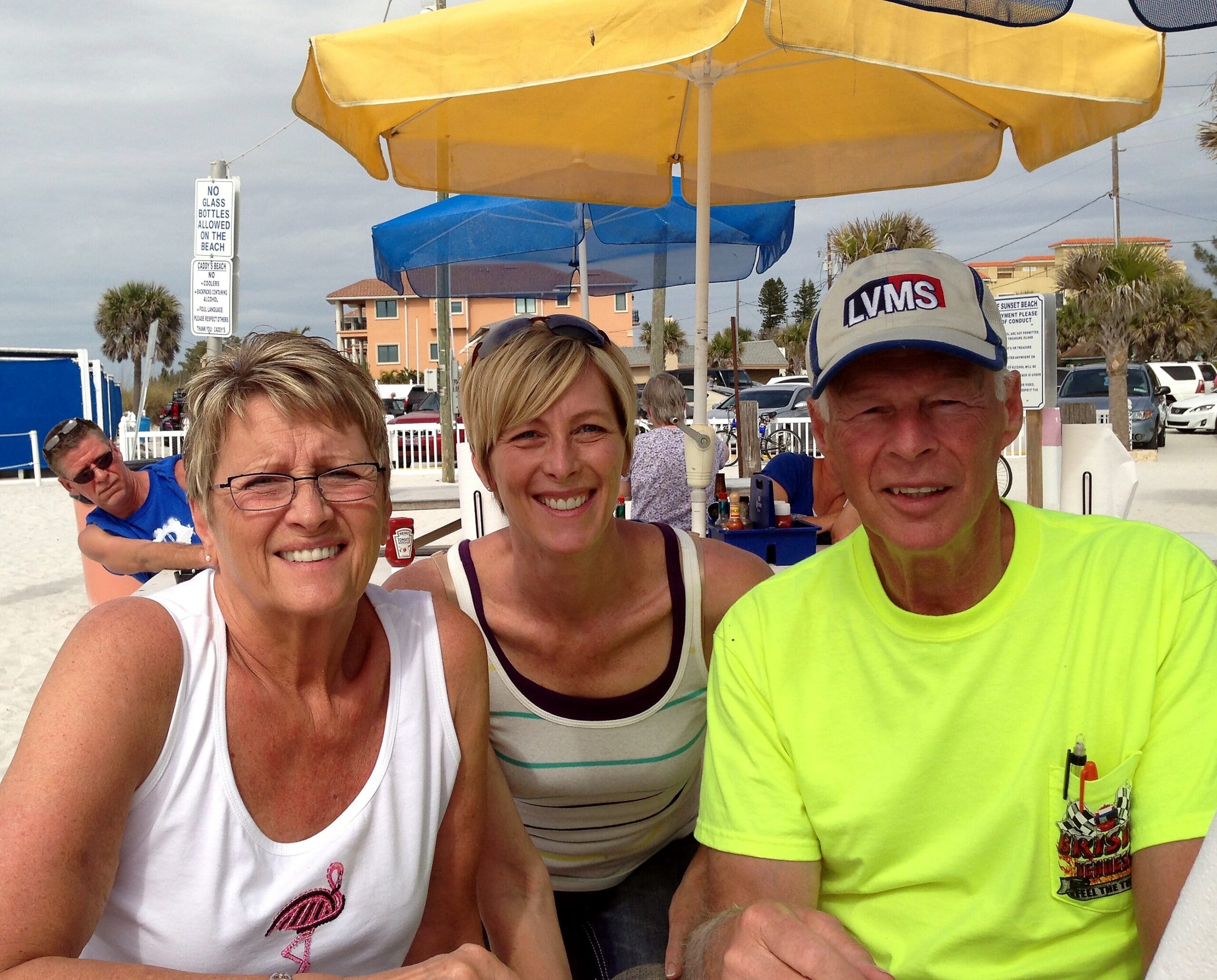 Group of smiling adults taking a beachside selfie under yellow and blue umbrellas with palm trees and parked cars in the background