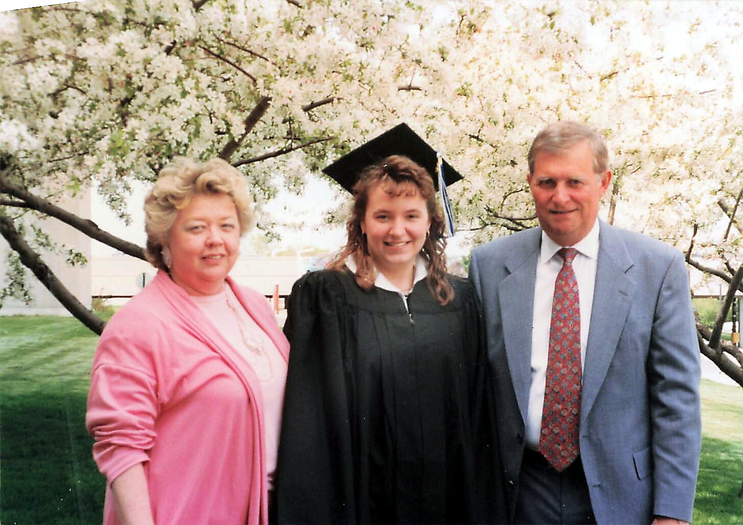 Graduate in cap and gown flanked by a woman in pink and a man in a blue suit outdoors under a flowering tree.