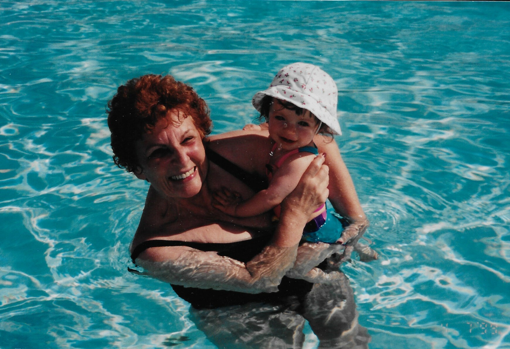 Smiling woman in a pool holding a young child wearing a white hat with small patterns.
