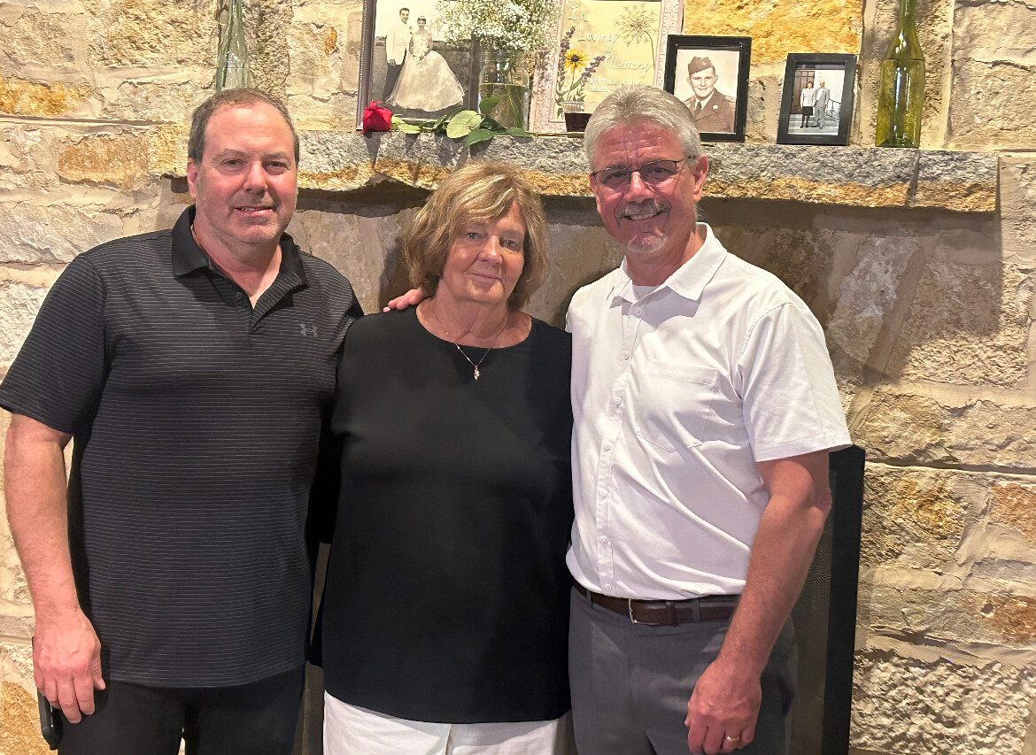 Three adults stand together and smile in front of a stone fireplace with framed photos on the mantel.