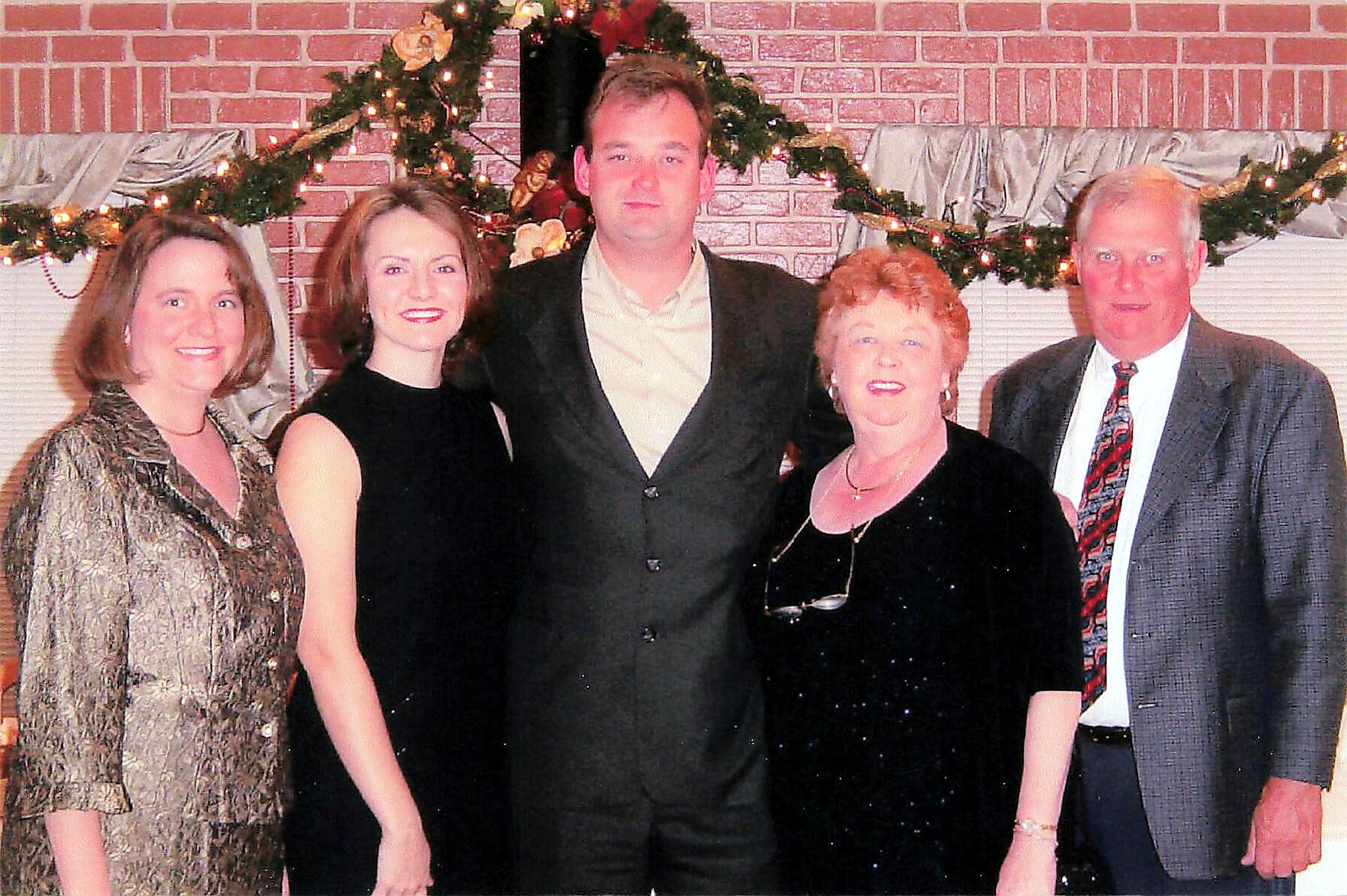 Five adults posing together at a holiday gathering in front of a brick wall with garlands and lights.