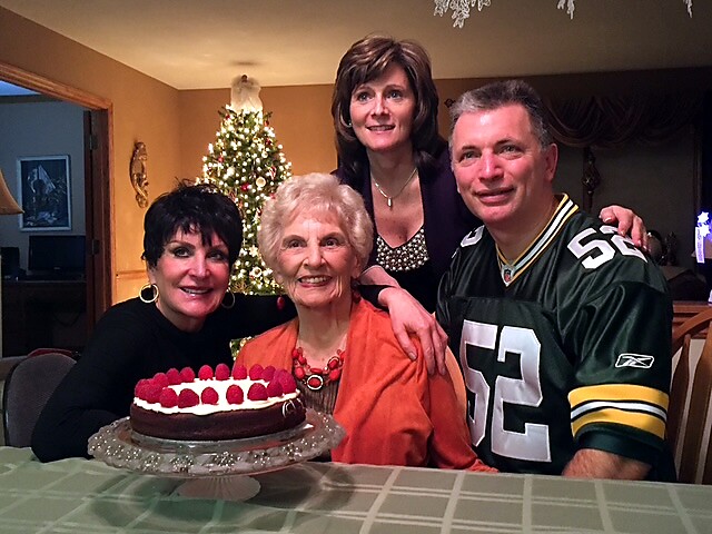 Four adults smiling around a birthday cake on a table, with a Christmas tree in a warm home setting.