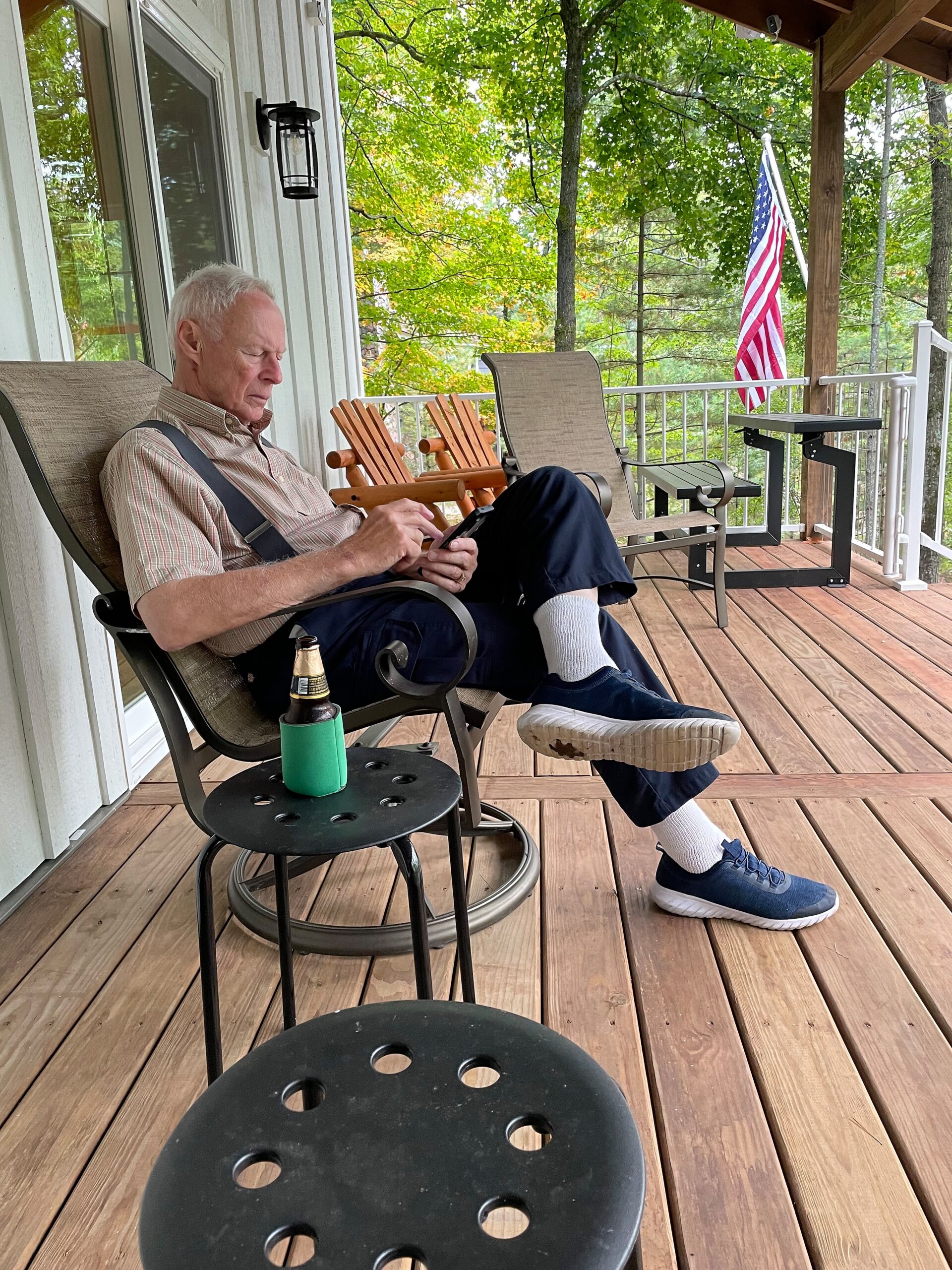 Older man in suspenders sits on a wooden porch chair, looking at a smartphone; a beer bottle in a green koozie rests on a nearby table, with an American flag on the railing and green trees in the background.