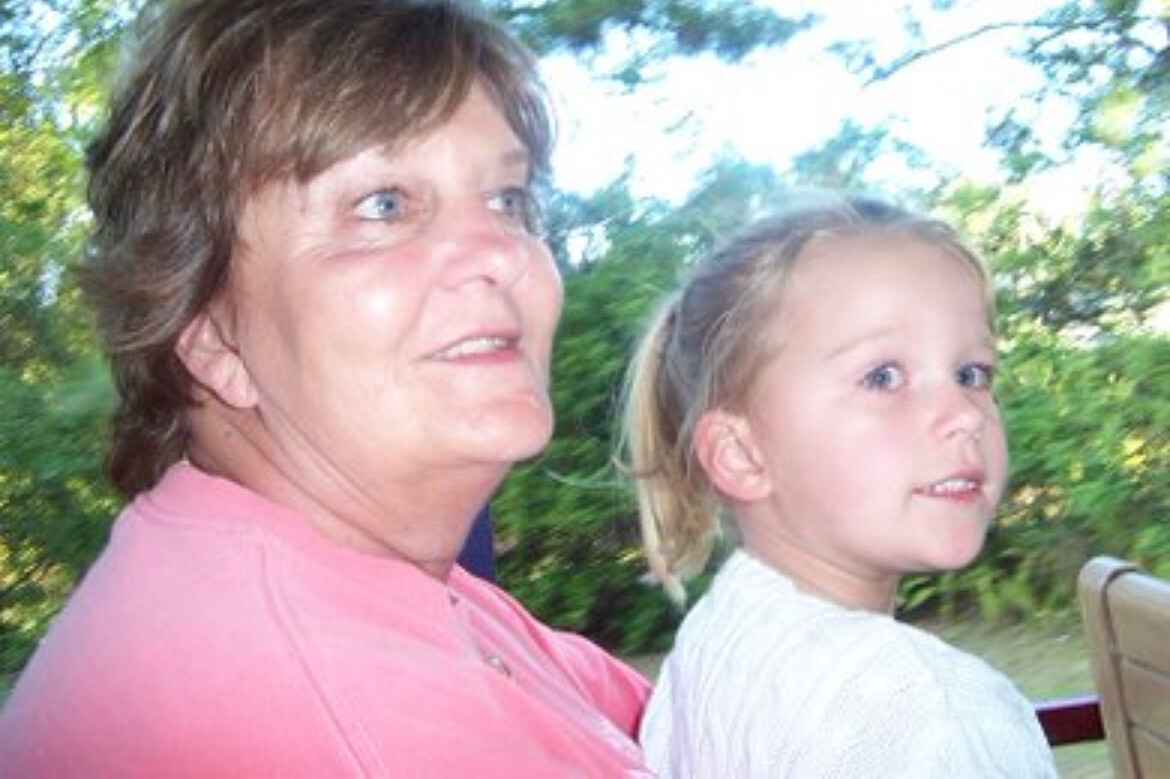 Woman in a pink shirt sits beside a young girl with blond hair in a ponytail outdoors, both looking to the right with greenery in the background.