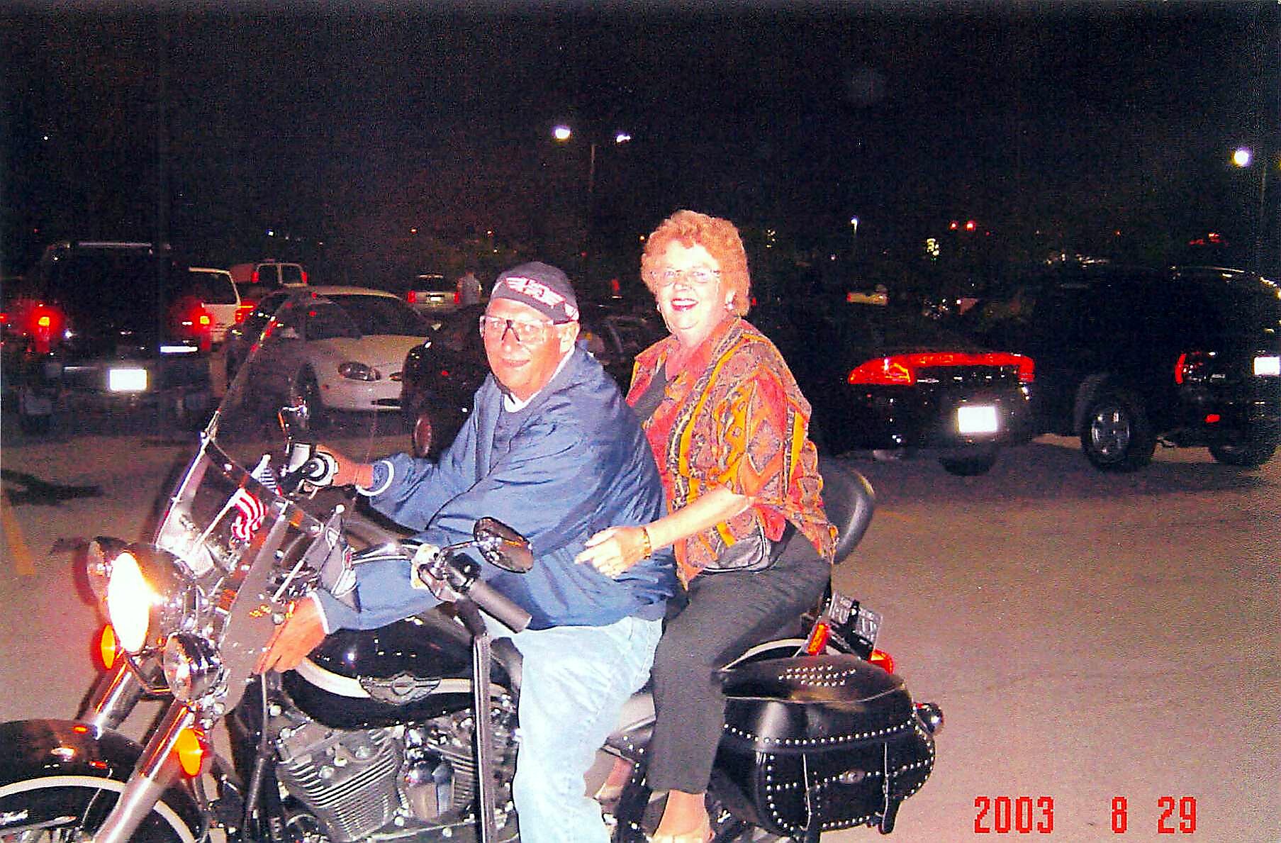 Man and woman ride a motorcycle at night in a parking lot; rider wears a bandana and glasses, passenger in a colorful blouse smiles.