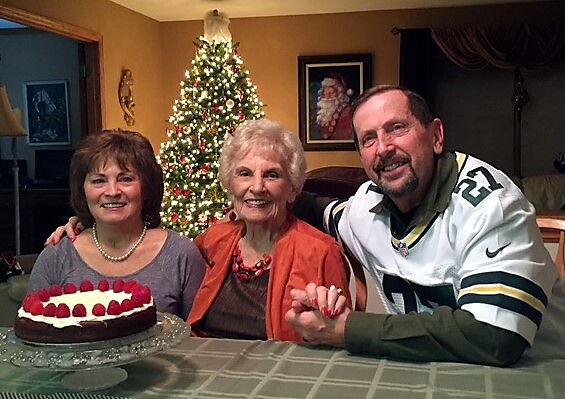 Three people sit at a dining table with a chocolate cake topped with raspberries, in a festively decorated room.