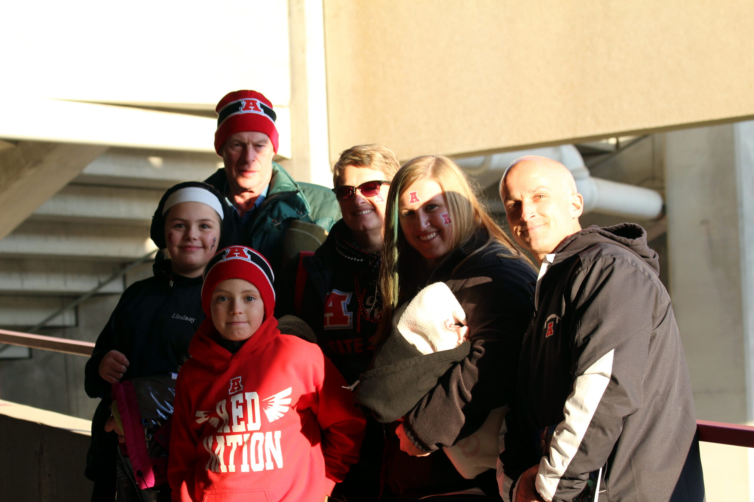 Group of six people posing outdoors in winter jackets, several wearing red hats with A logos.
