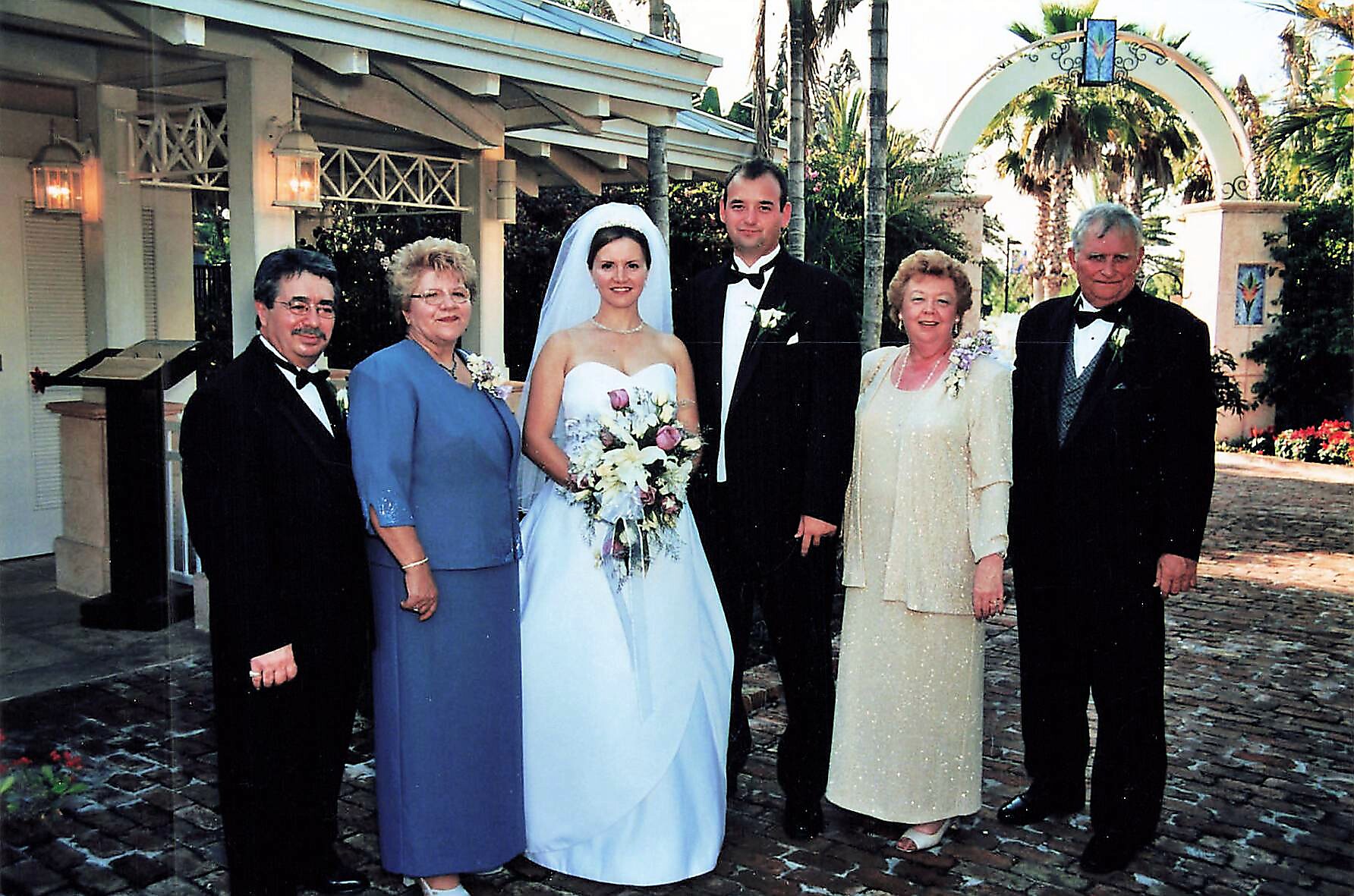 Bride in a white gown with veil and bouquet, flanked by the groom and four family members outdoors at a wedding venue.