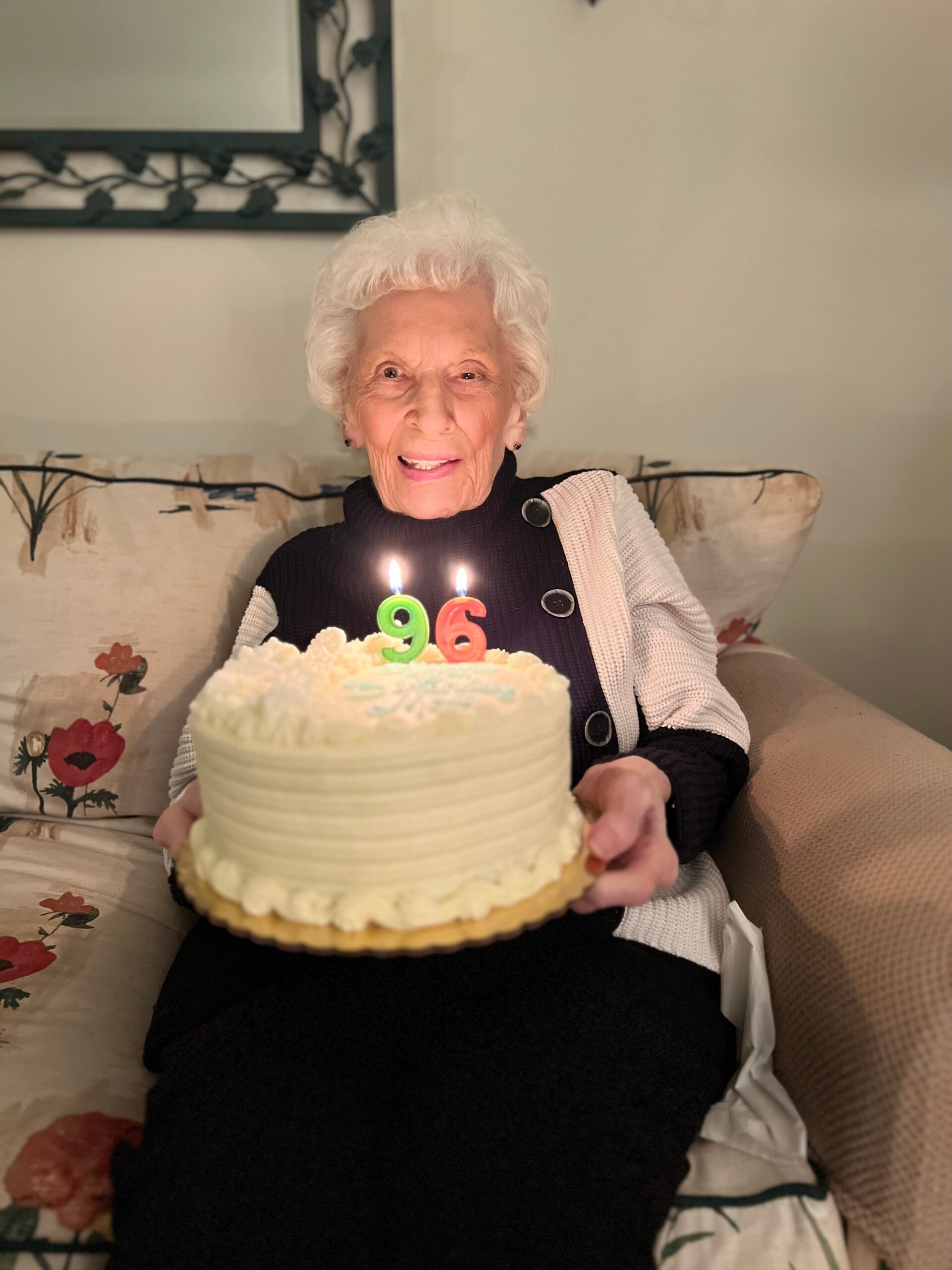 Smiling elderly woman sitting on a patterned sofa, holding a frosted birthday cake with lit candles showing 9 and 6 (96).