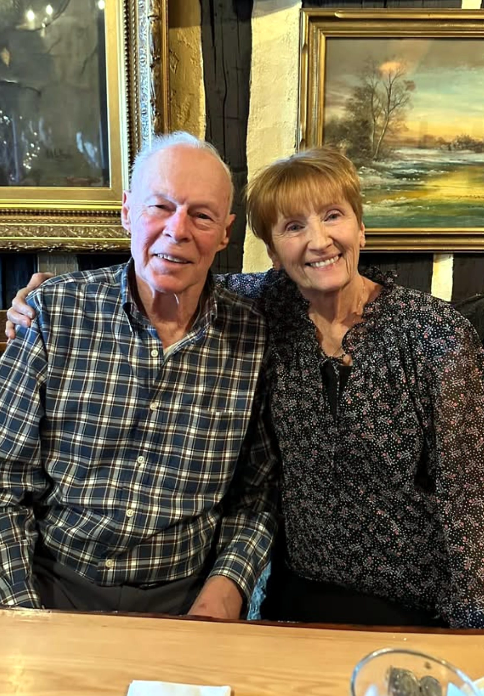 Smiling elderly man and woman sitting together at a restaurant table with framed paintings behind them.