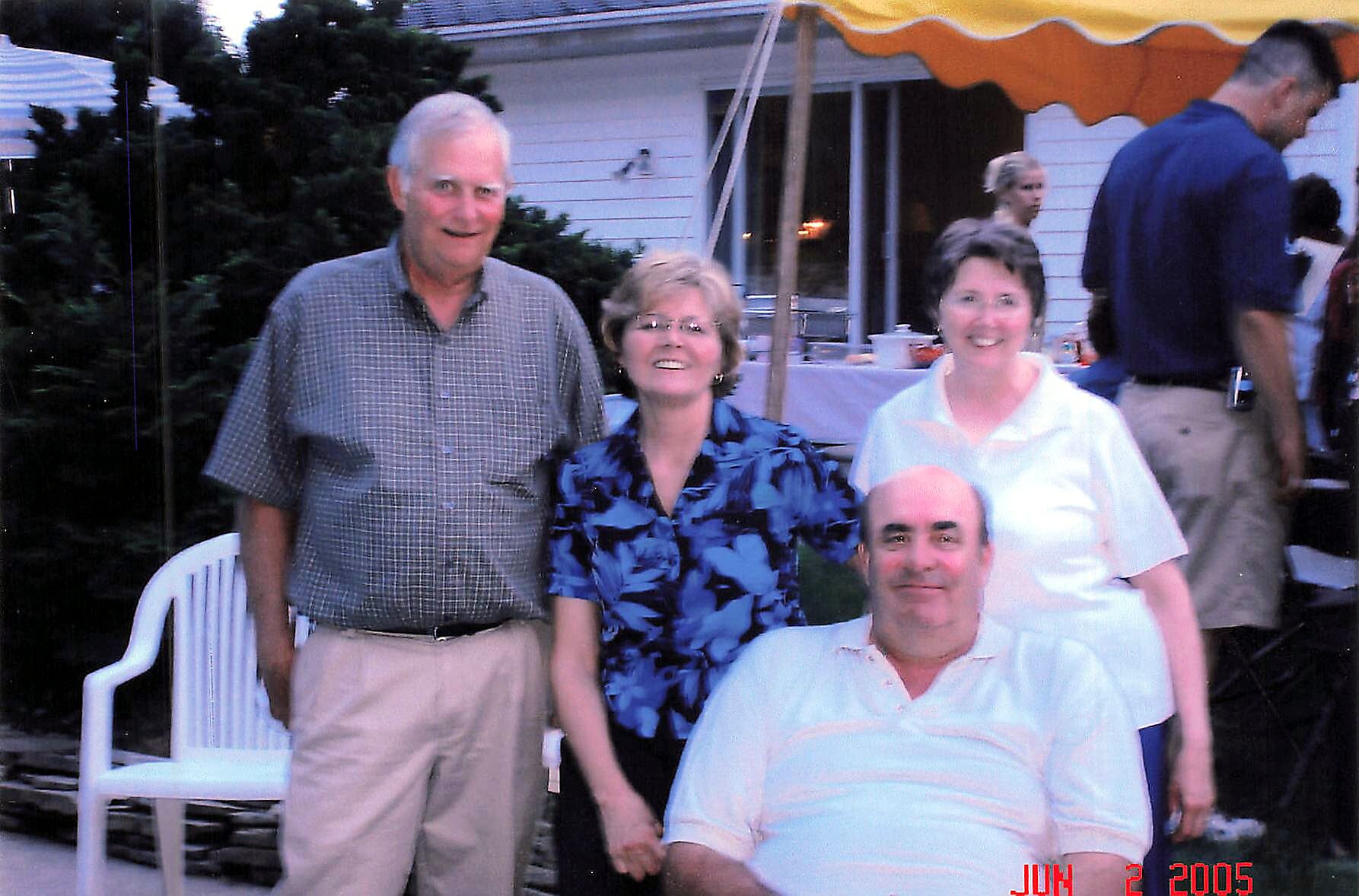 Group of four adults posing in a backyard at a casual gathering, two women in the center and two men on the sides, under a yellow canopy. Date stamp reads JUL 2 2005.