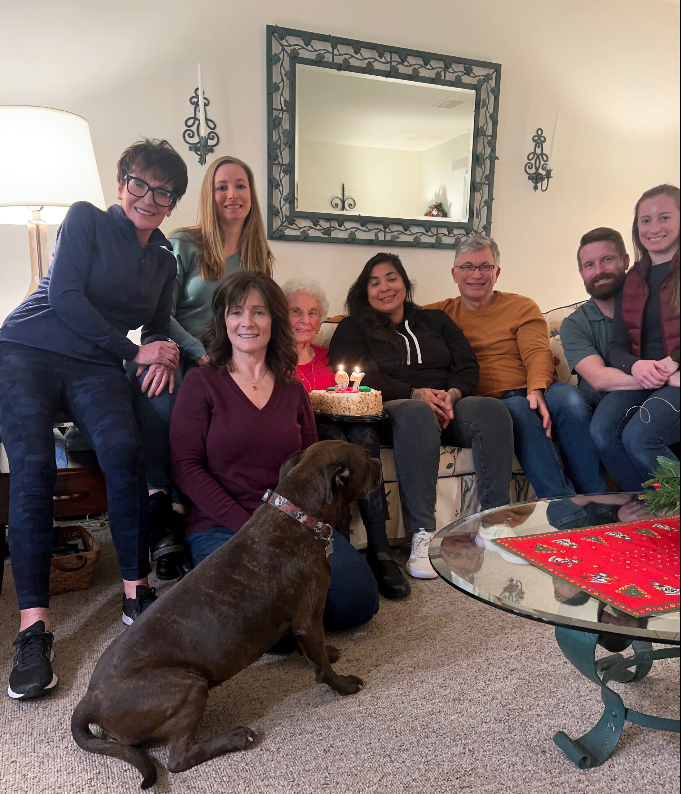 A group of adults seated on a couch in a living room, smiling around a birthday cake with lit candles, with a dark dog in the foreground on the carpet.