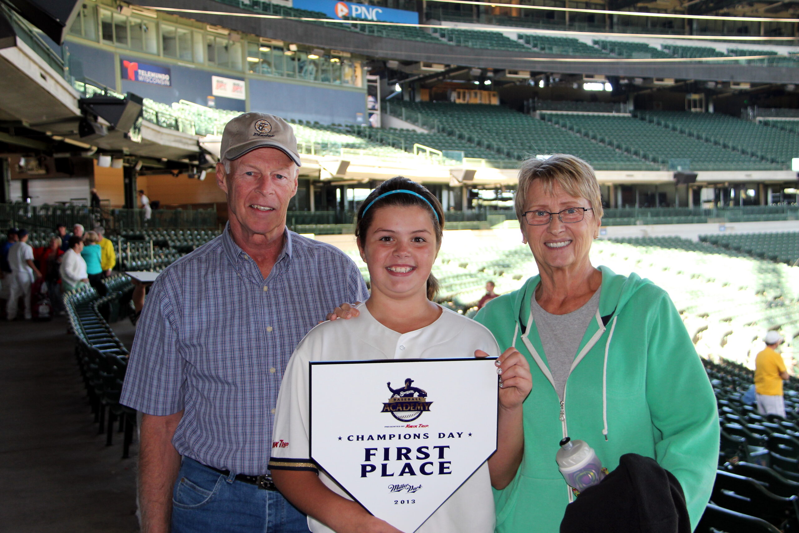 Girl in a white sports uniform holding a 'First Place' plaque, flanked by an older man and woman in a baseball stadium.