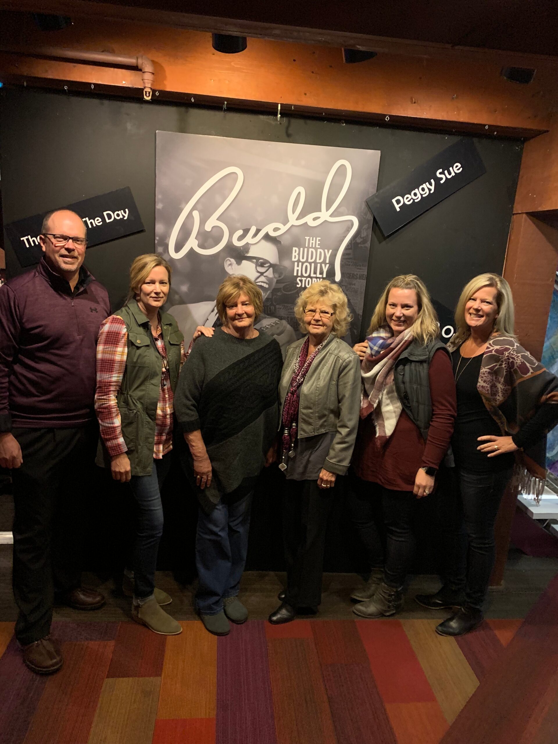 Seven adults pose together in front of a large Buddy Holly Story poster on a wall inside a venue, smiling for the photo.