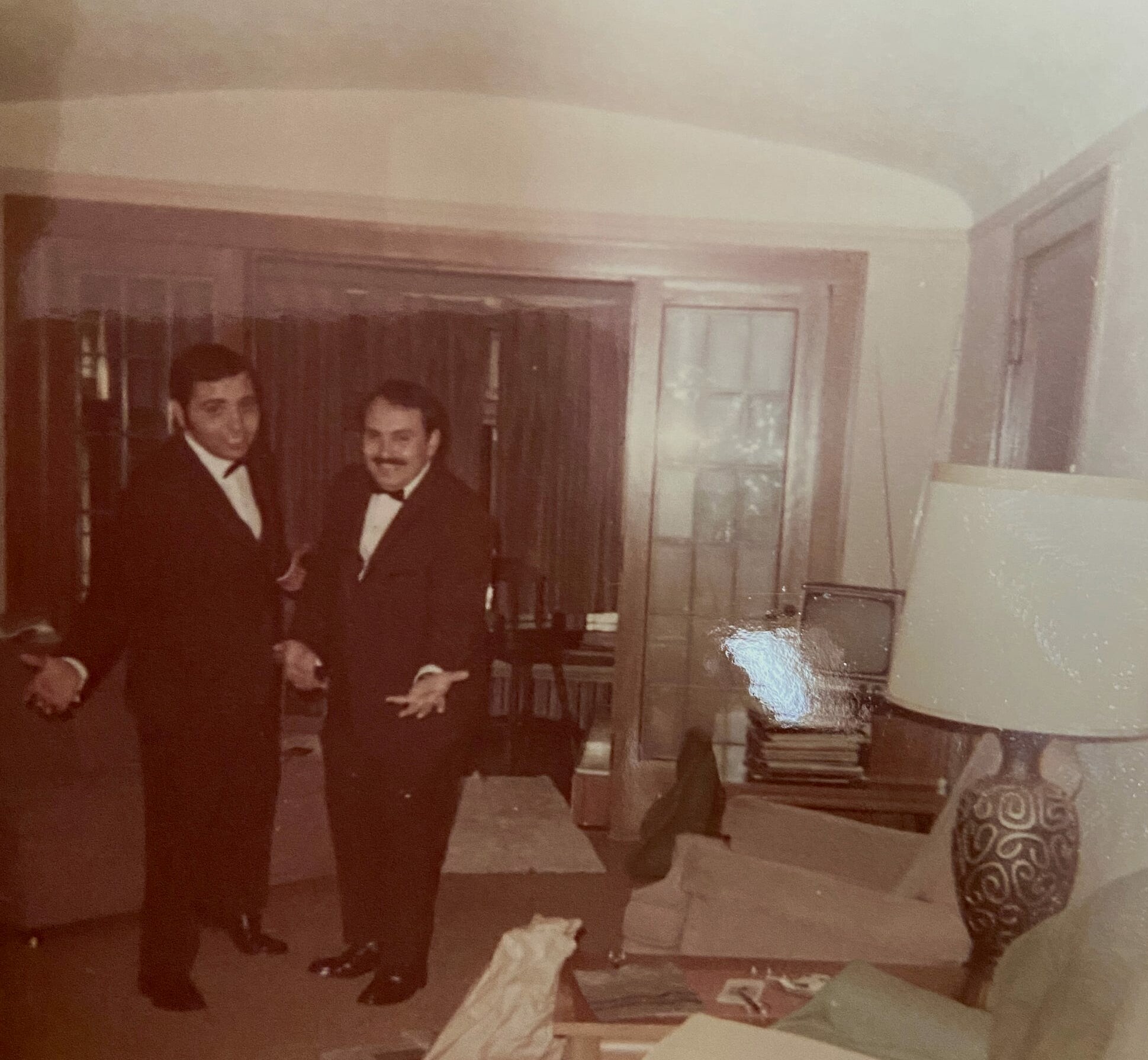Two men in tuxedos pose together in a sepia-toned living room with cabinets and a patterned chair nearby.