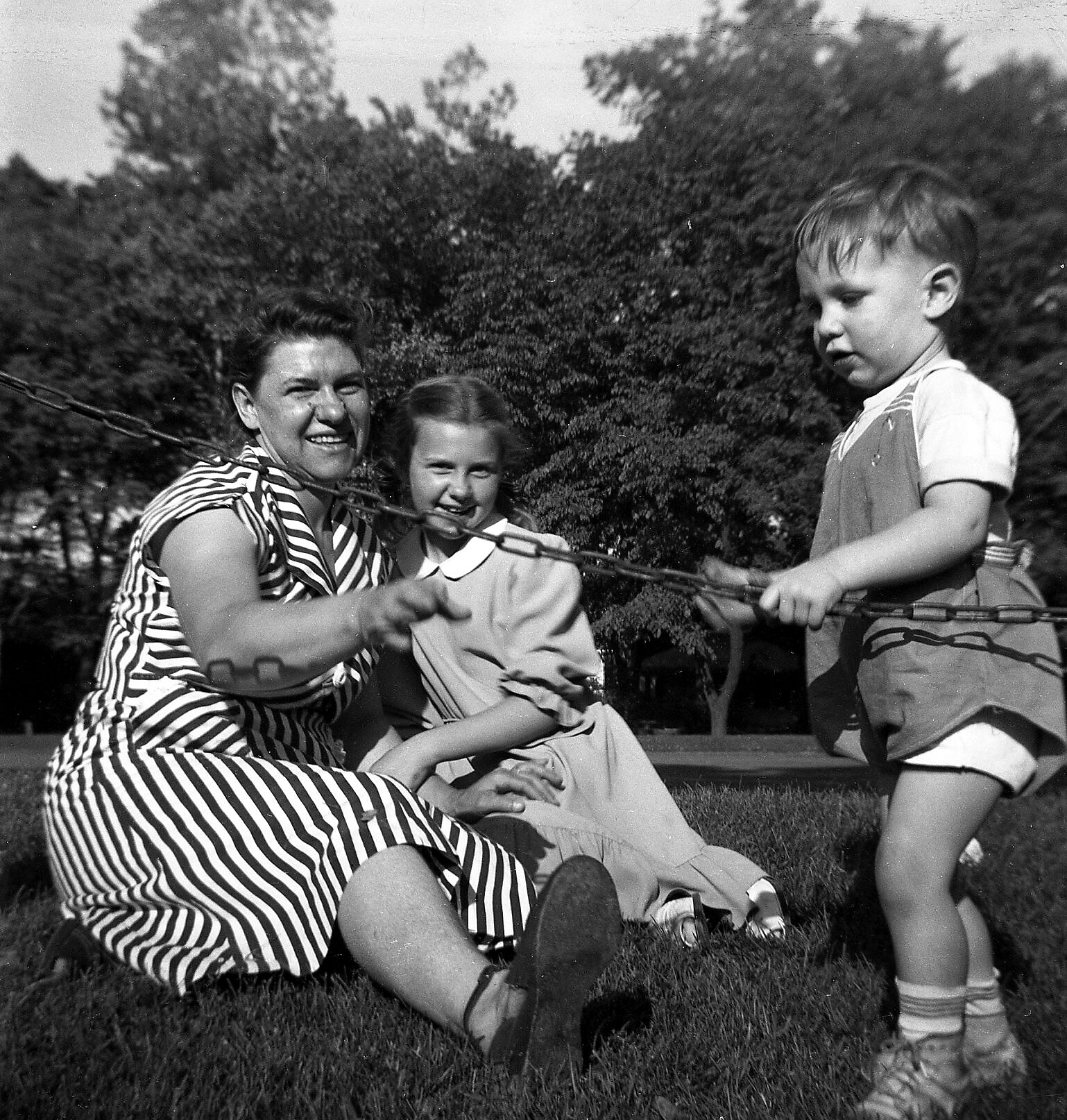 Woman in a striped dress sits on a swing as two children push it in a park.