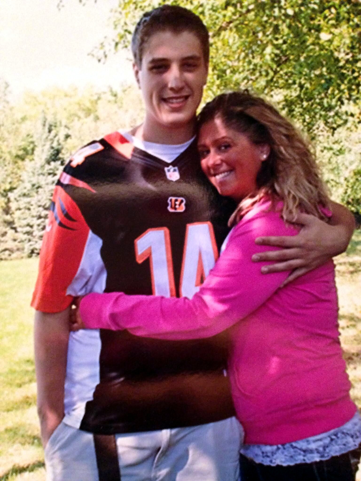 Smiling man in Cincinnati Bengals jersey (number 14) hugs a smiling woman in a pink top outdoors.