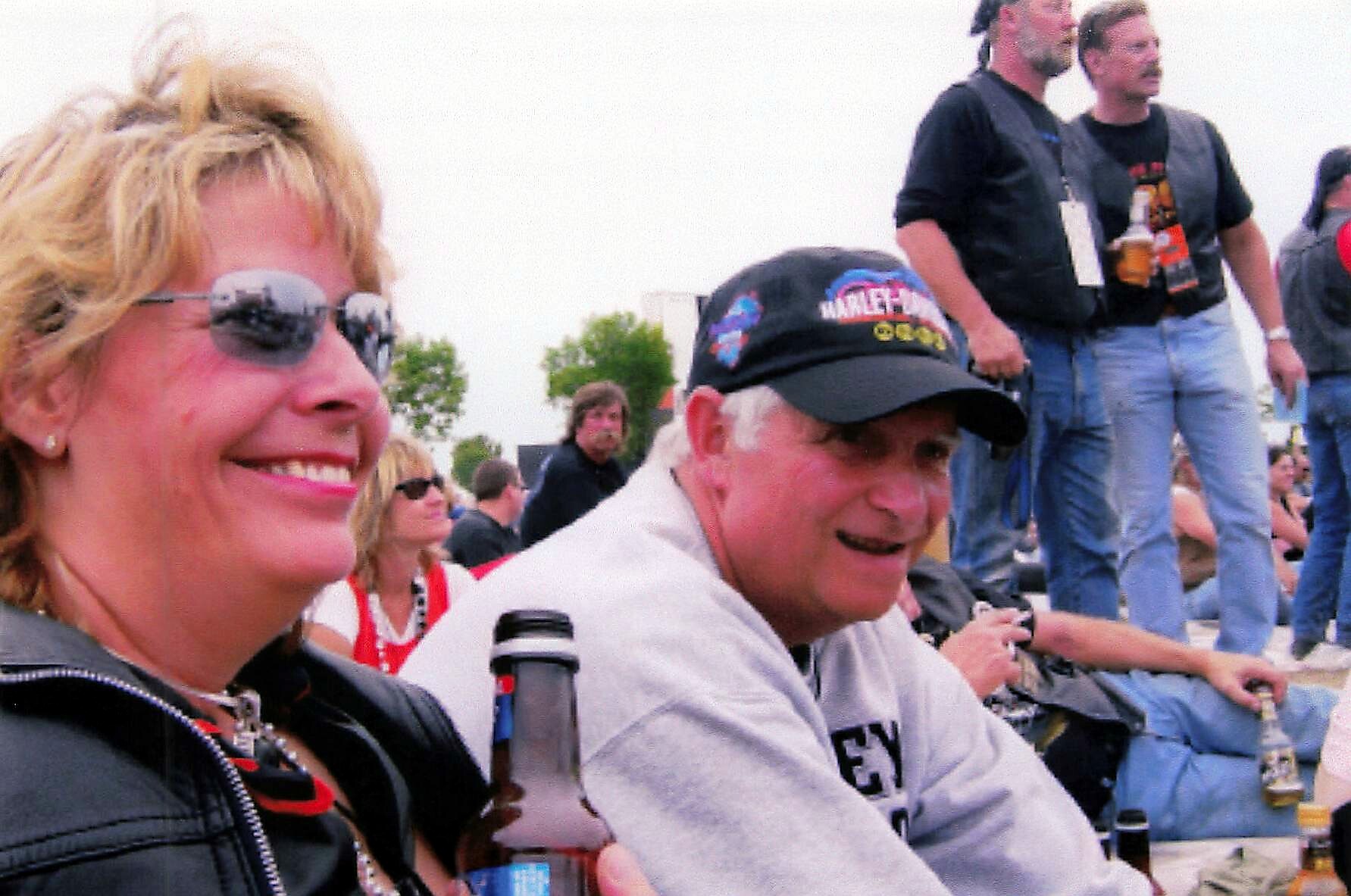 Smiling woman with blonde hair and sunglasses in the foreground, seated with others at an outdoor event; a man wearing a Harley-Davidson cap sits beside her, while people stand on bleachers in the background.