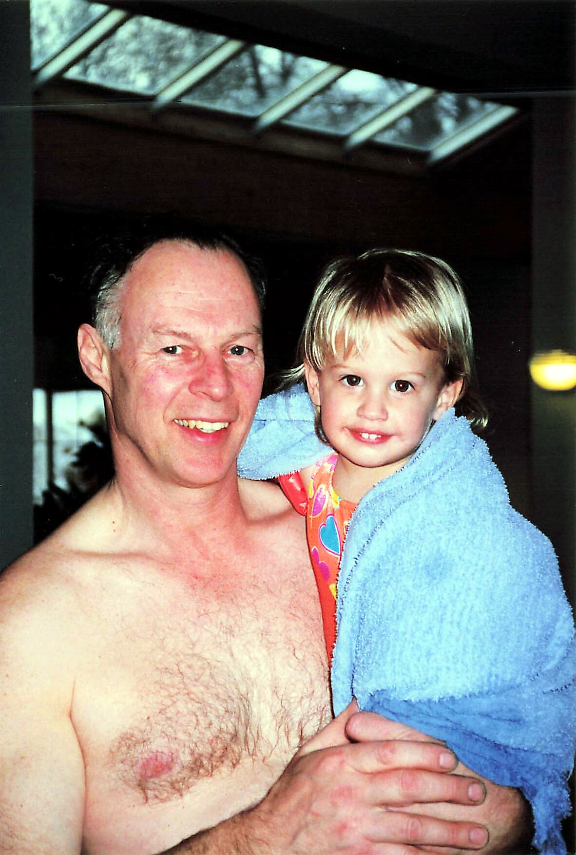 Smiling man with short gray hair holding a young girl wrapped in a blue towel indoors, both looking at the camera.