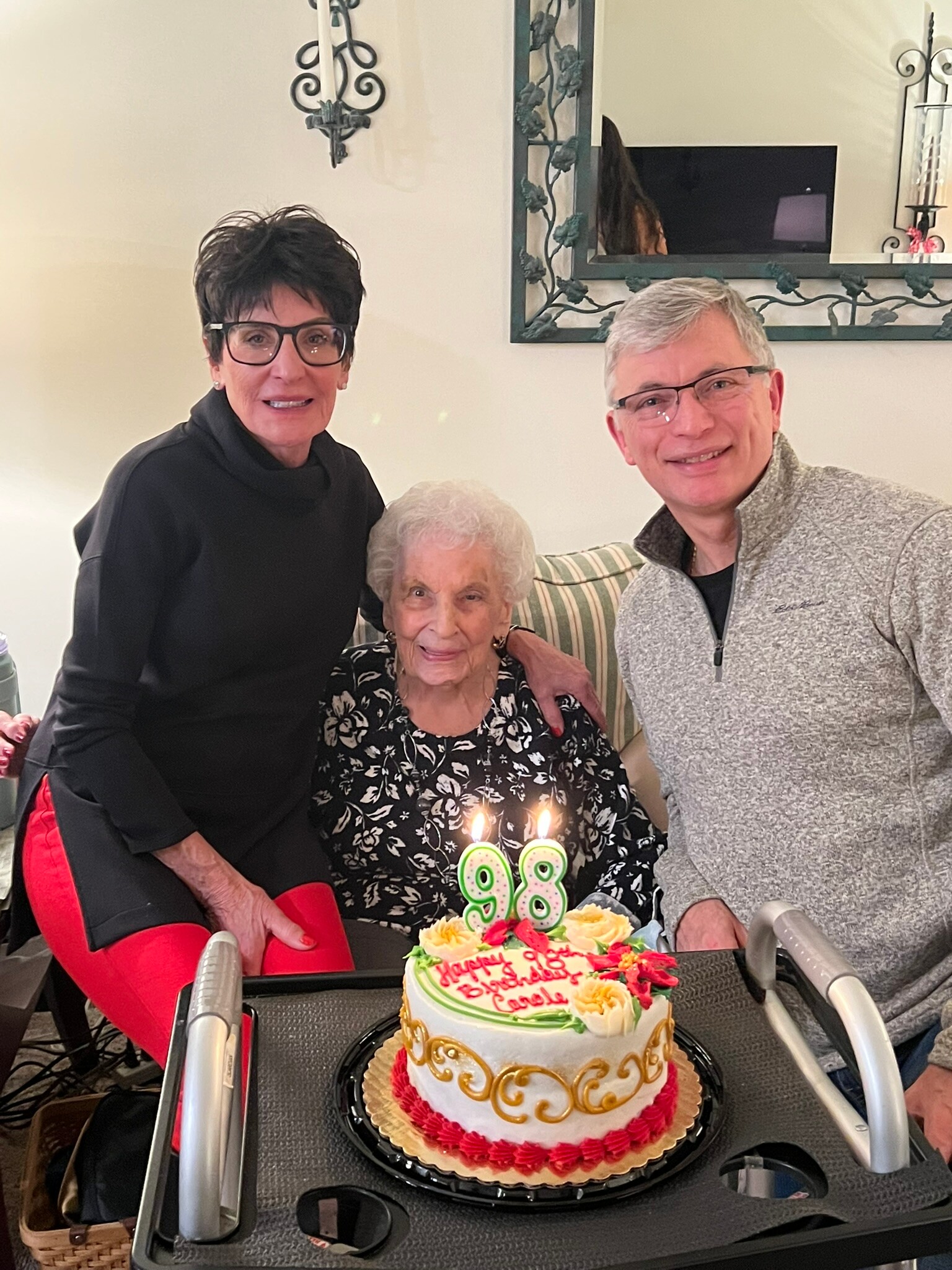 Three people celebrate a birthday around an elderly woman in a floral dress; a cake with green and yellow decorations and glowing '98' candles sits on a table in front of them.