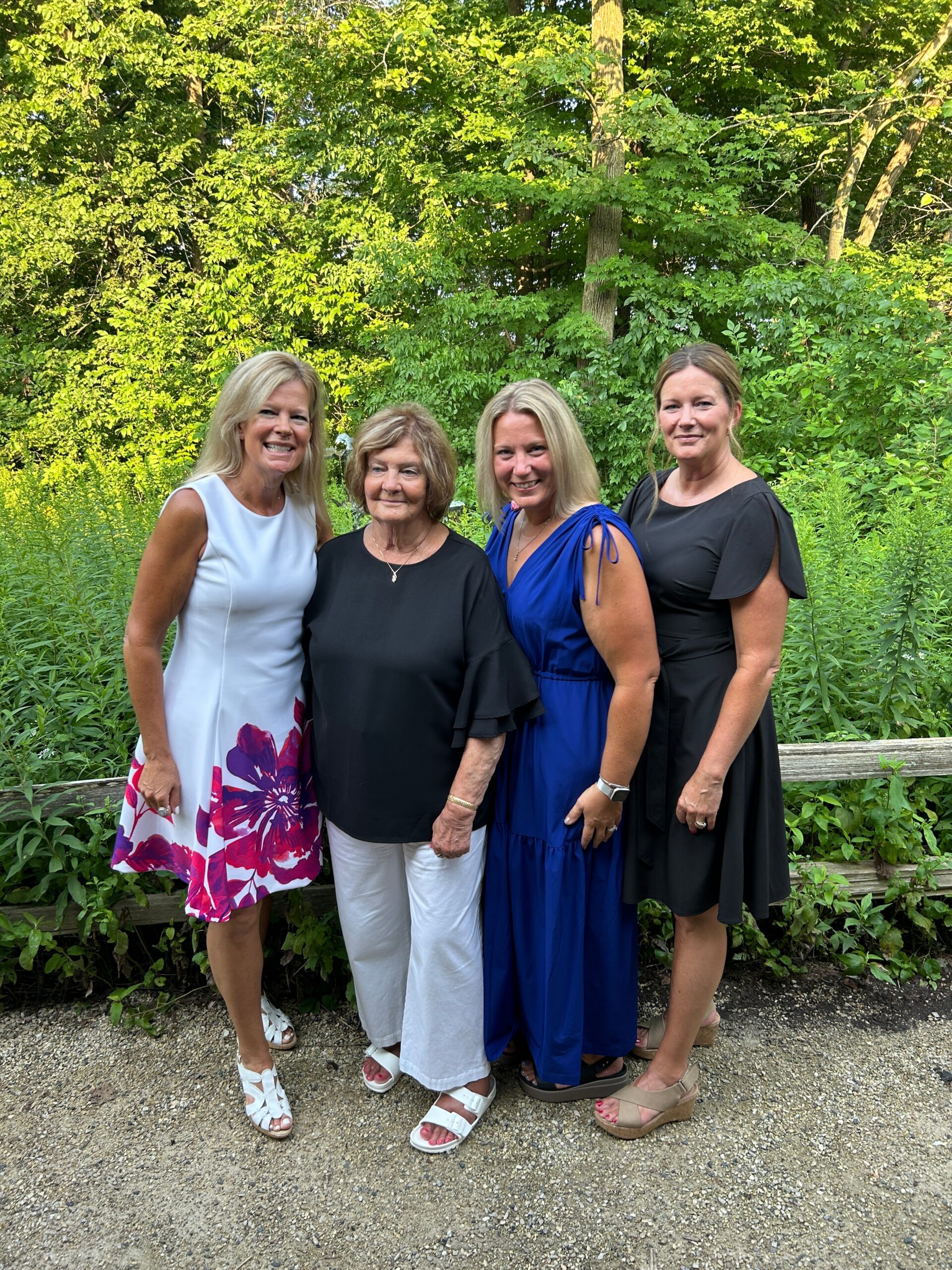 Four women posing together outdoors in a garden, smiling, wearing white, black, blue, and black dresses.