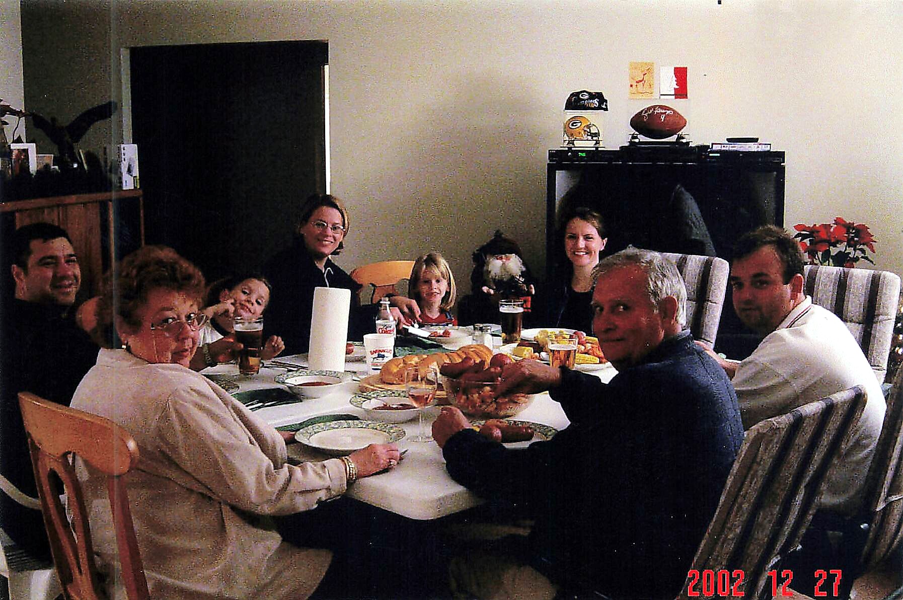 A family gathering around a dining table for a holiday meal, with plates, bread, and drinks, smiling at the camera; date stamp 2002-12-27 visible in the corner.