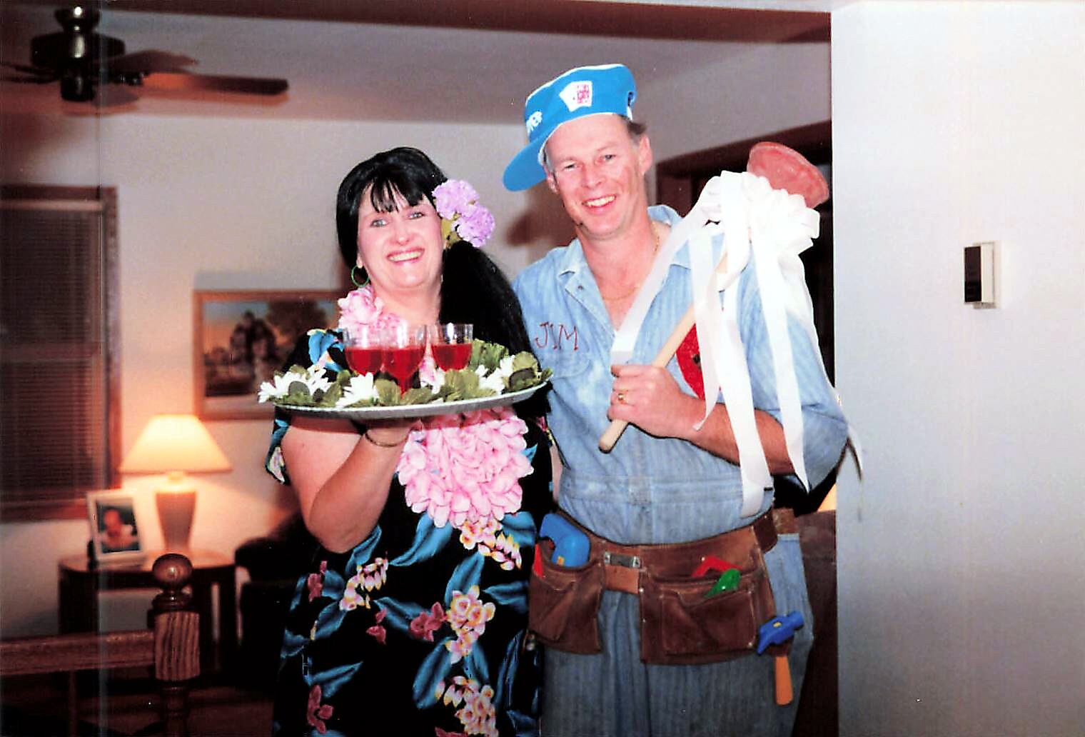 Smiling woman in a floral dress with a pink flower lei holds a tray of red drinks garnished with greenery; beside her, a man in a blue shirt and cap smiles, holding white ribbons and a prop.