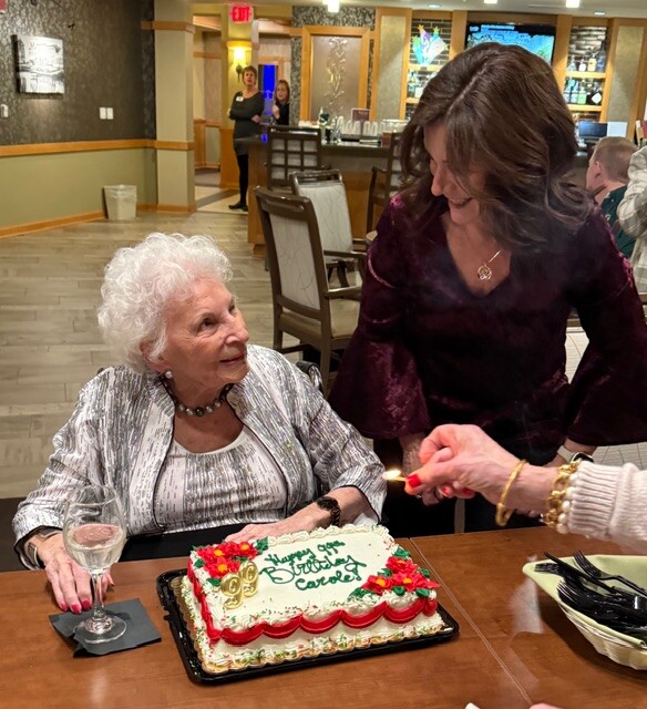 Older woman with white curls sits at a table with a birthday cake reading 'Happy Birthday Carol'; a younger woman lights a candle nearby.