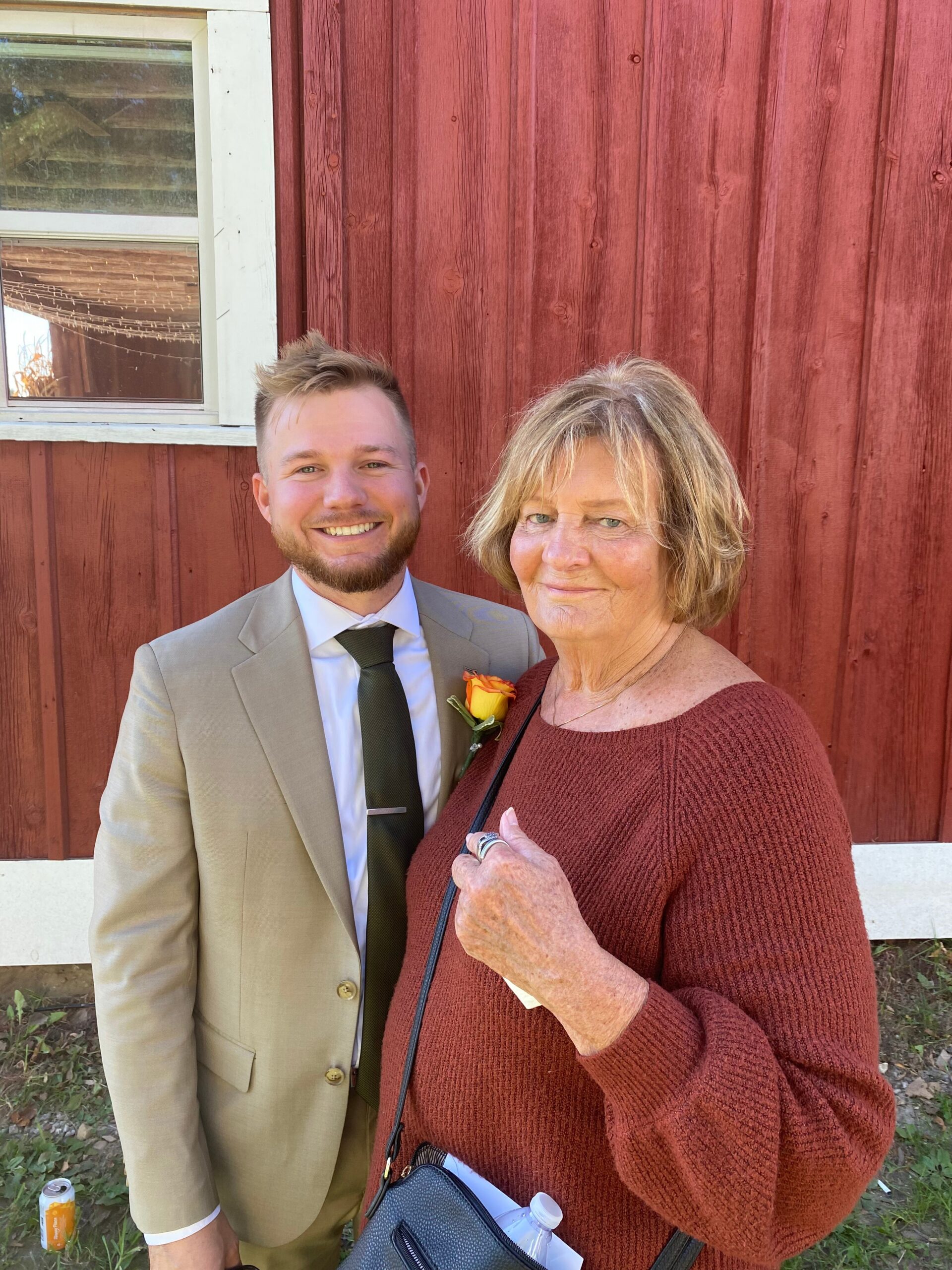 Smiling man in a beige suit with a boutonniere stands beside an older woman in a maroon sweater, posing outside against a red wooden wall.