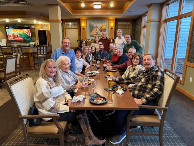 Group of about 20 people sitting around a long dining table in a warmly lit restaurant, sharing a meal and drinks while smiling at the camera.
