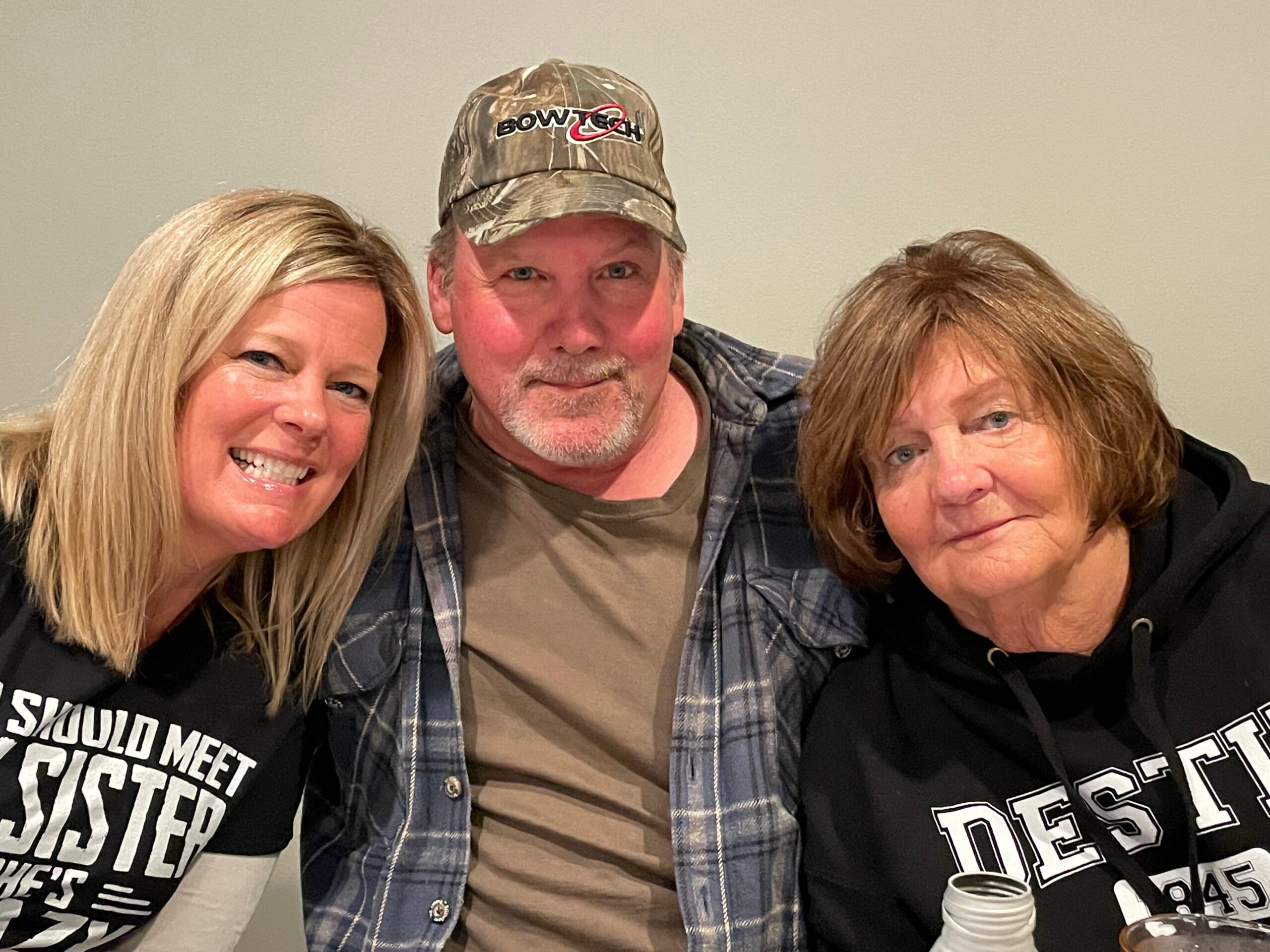 Three adults posing closely for a casual group photo, smiling at the camera; man in the middle wears a camo cap and plaid shirt, flanked by two women in black shirts with bold white text on them.