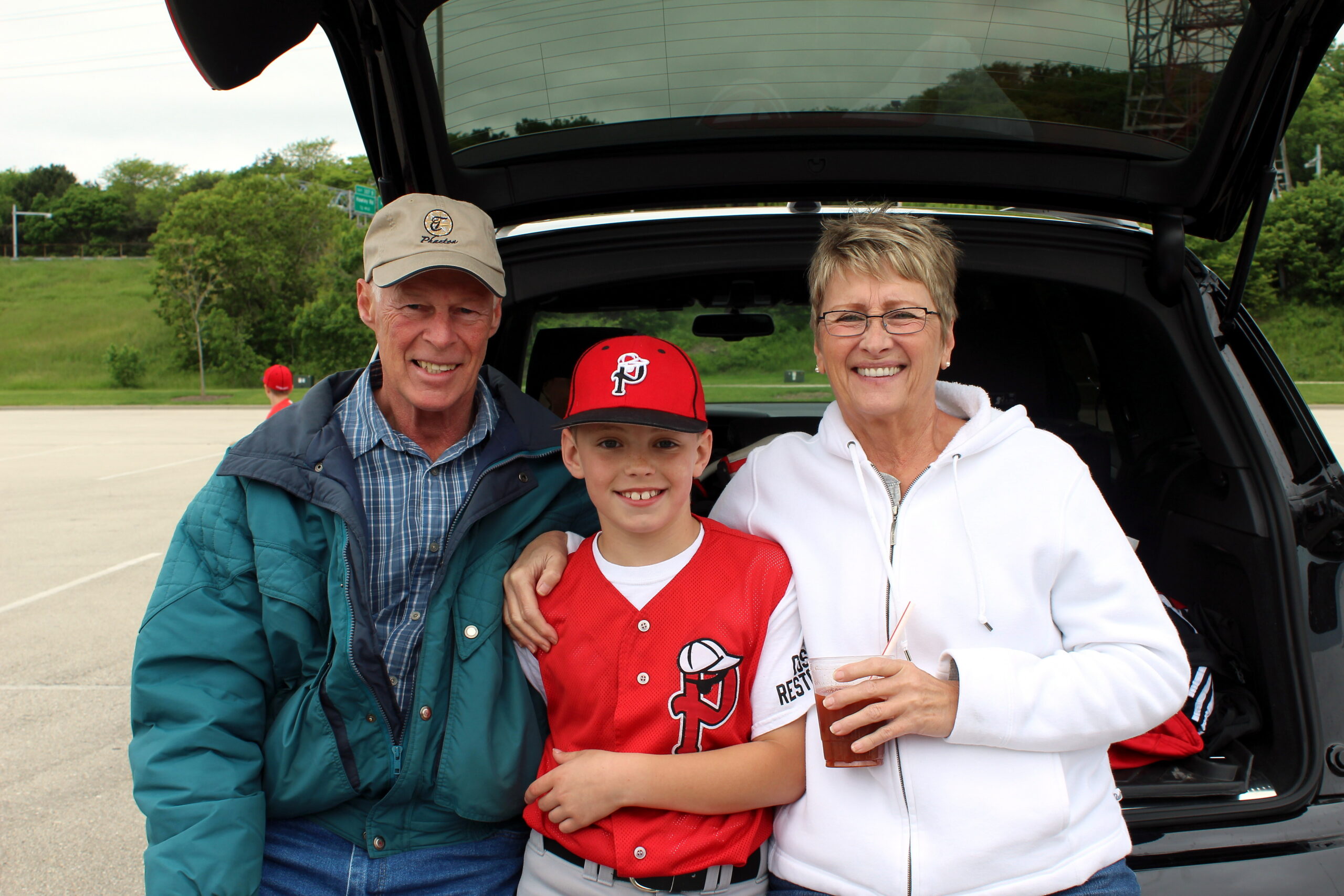 Family of three smiling in a car trunk outdoors after a game, man in cap, boy in red jersey, woman in white hoodie with drink