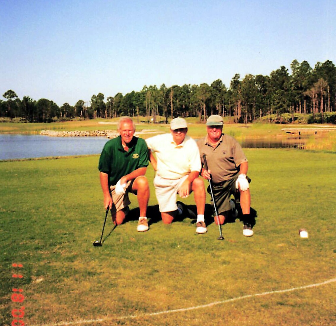 Three men kneel on a golf course green with clubs, posing for a photo near a water hazard and trees in the background.