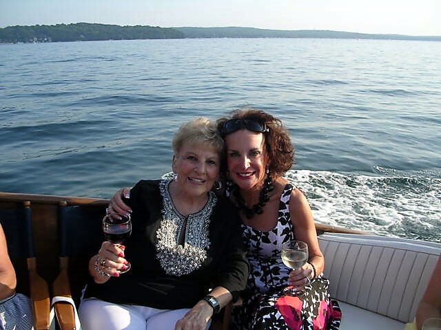 Two women on a boat, arm in arm, smiling, each holding a glass of wine with water and shoreline in the background.