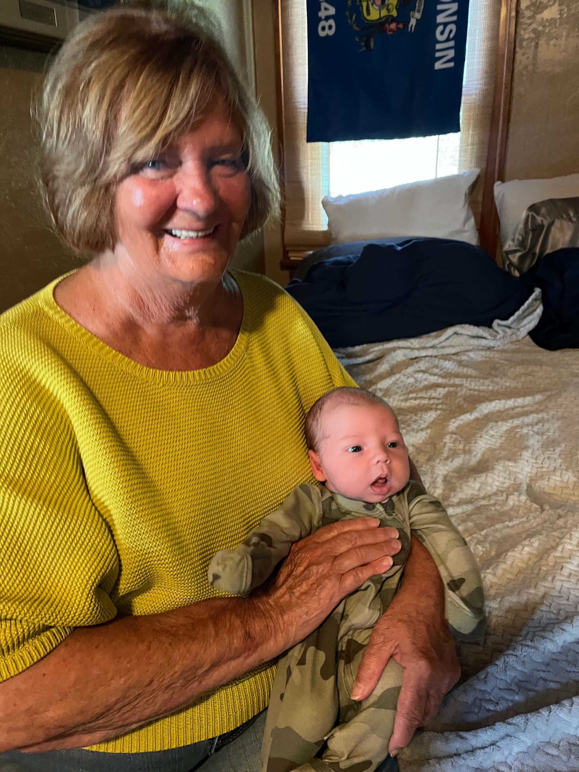 An elderly woman in a yellow knit sweater holding a newborn baby on a bed in a cozy bedroom.