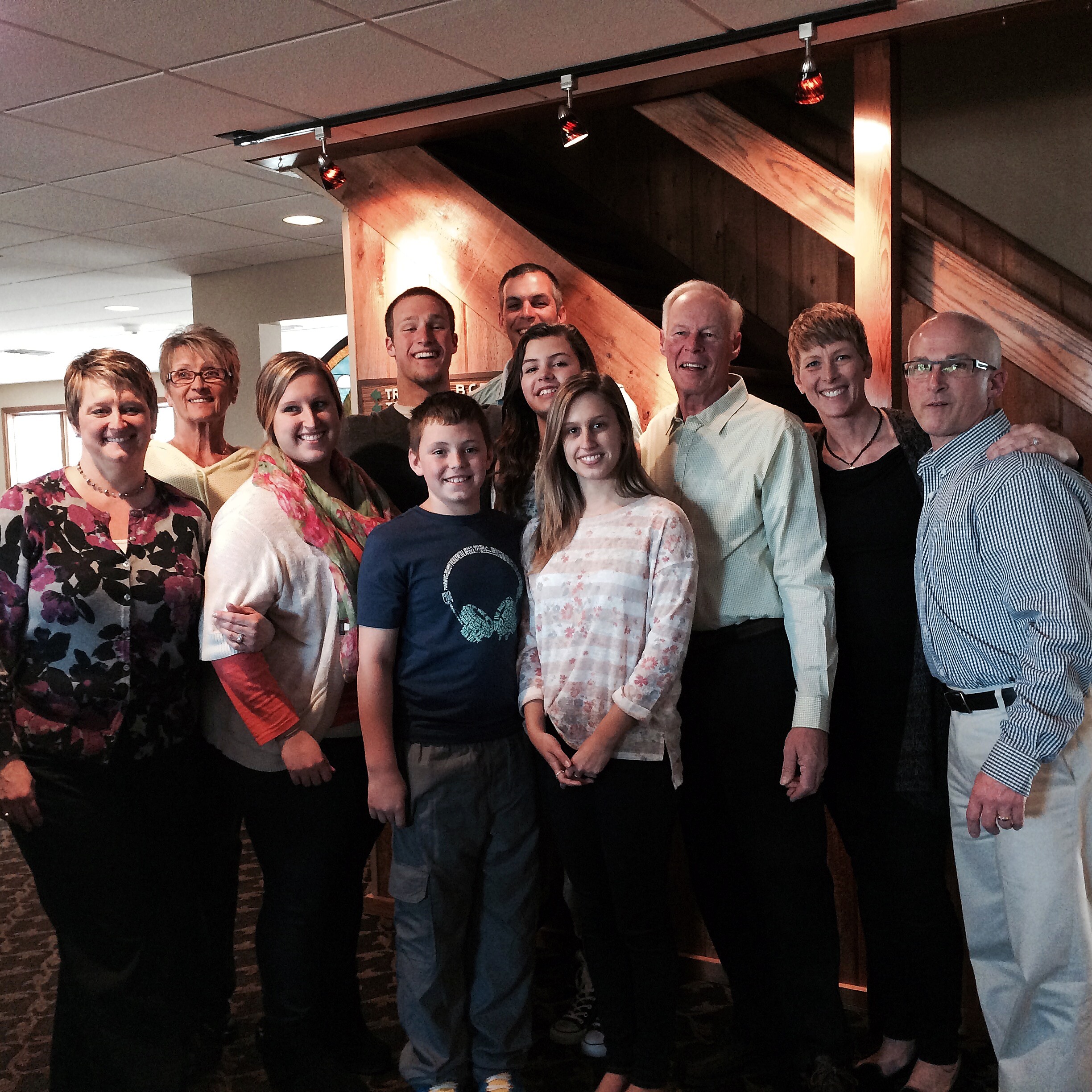 Group of people of different ages posing closely together indoors, smiling, with wood paneling and warm lighting in the background.