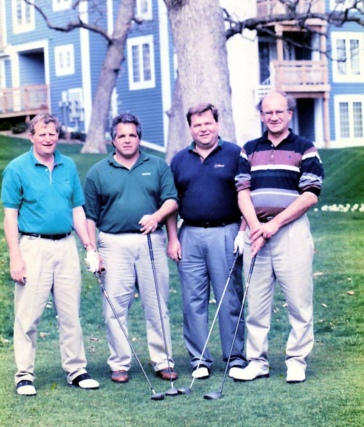 Four men posing on a golf course with their clubs, standing on the green grass in front of blue buildings in the background.