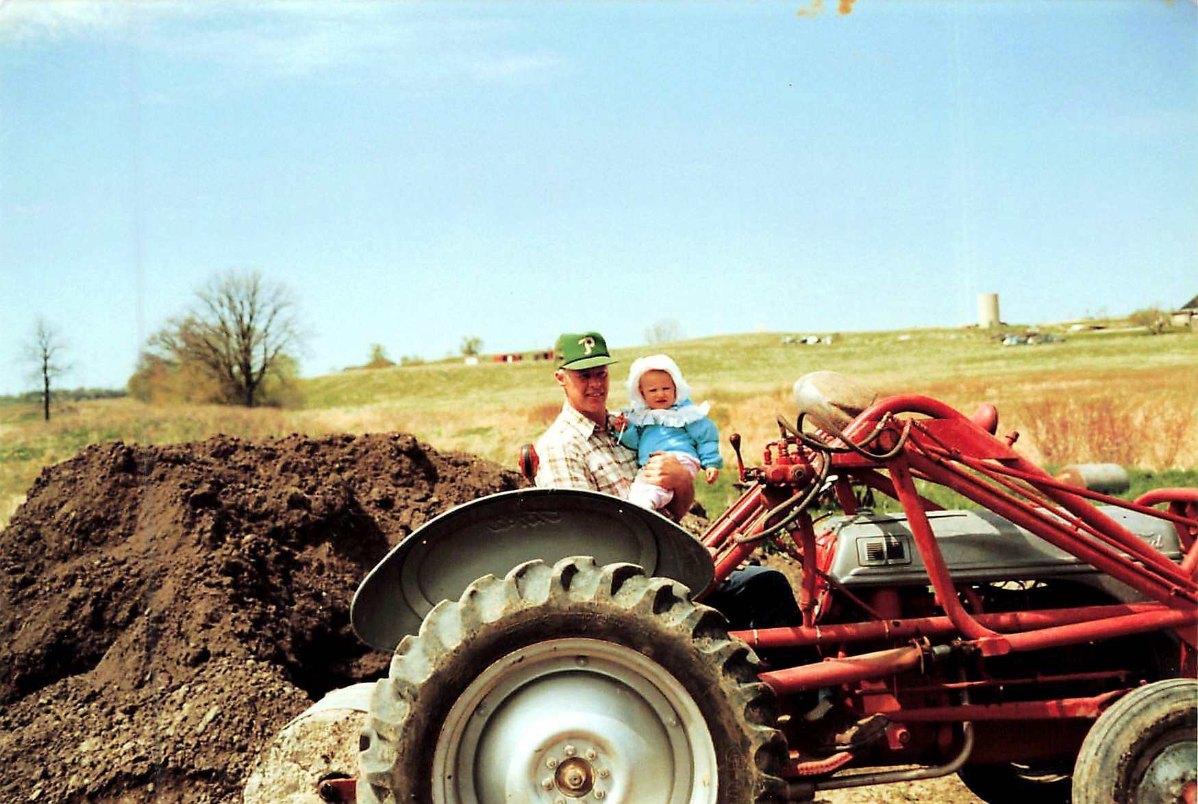 Man wearing a green cap and plaid shirt holds a baby in a blue outfit while seated on a red tractor next to a large dirt pile in a rural field on a sunny day.