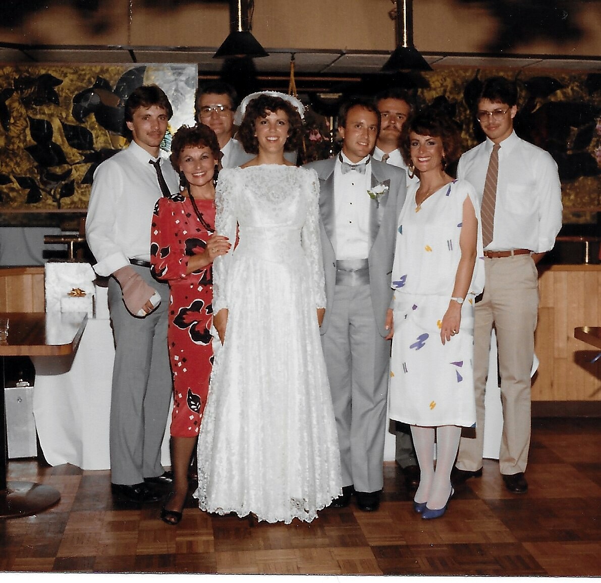 Bride in a lace wedding gown and groom in a gray suit pose with family at a wedding reception.